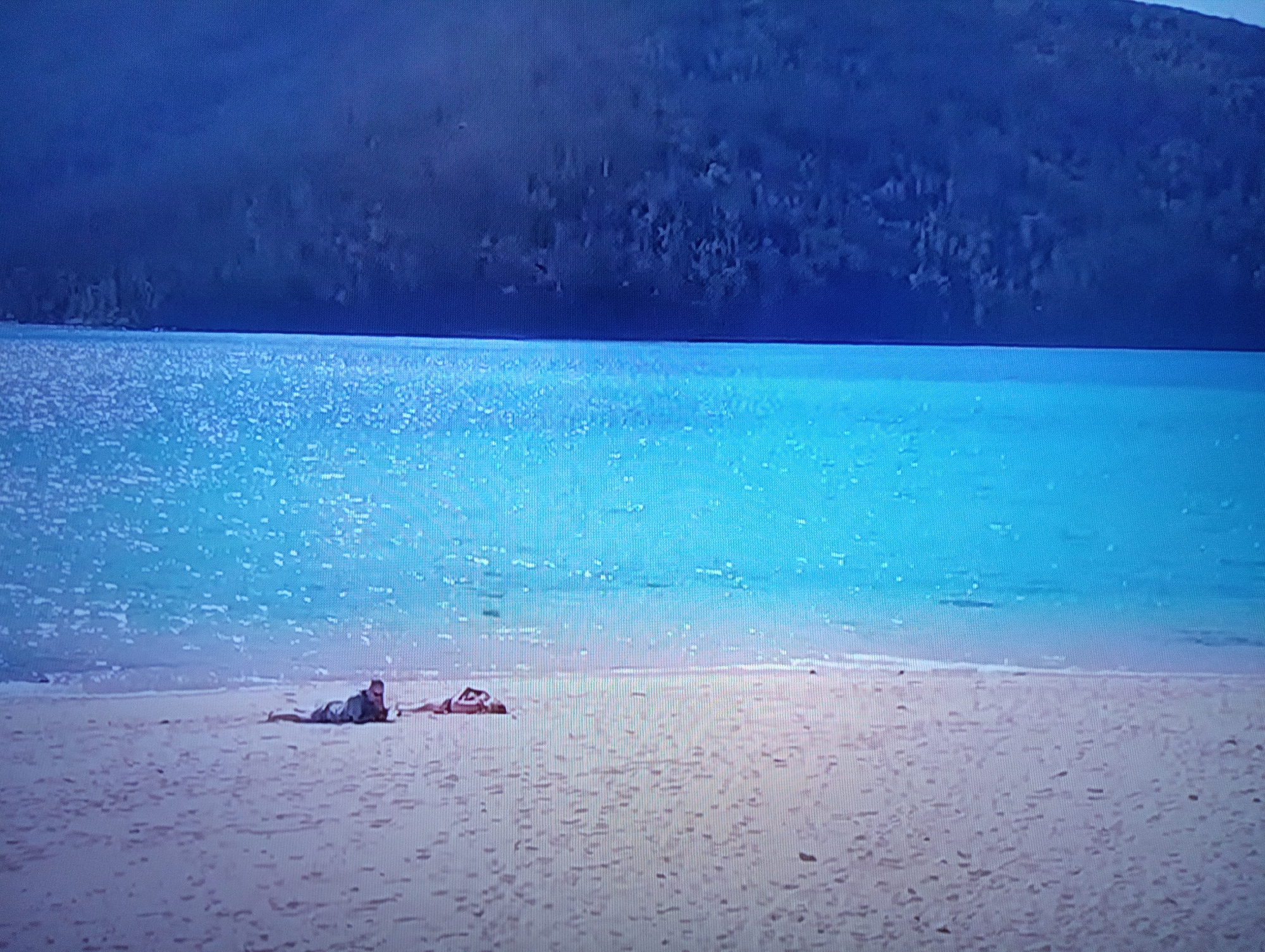 The image displays a serene beach scene with a person lying on the sand near the water's edge. The environment features a vast expanse of clear, blue water meeting a sandy beach. In the background, a dark, mountainous landscape is visible under a dusky sky. The person is lying on their stomach, partially obscured by their clothing, and appears to be resting or sunbathing. Another figure, possibly another person, is lying on the sand to the right of the main subject. The overall impression is one of tranquility and isolation in a natural setting. There is no information to suggest this scene is located in the city of Dnipro, Ukraine.