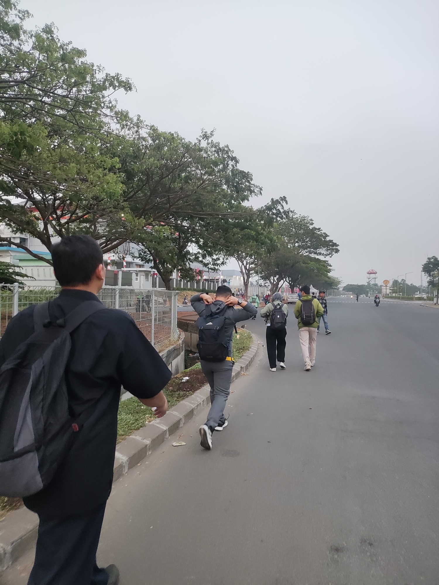 In Telagamurni, Indonesia, a group of people are walking along a paved road, likely for exercise or commuting. The sky is overcast, suggesting either early morning or a cloudy day. The road is bordered by a concrete sidewalk and a drainage ditch. Lush green trees line the opposite side of the road, with some buildings and a large mushroom-shaped structure visible in the distance. The people are dressed casually, with several wearing backpacks, indicating they may be students or hikers. The overall atmosphere is one of everyday activity in an urban setting.