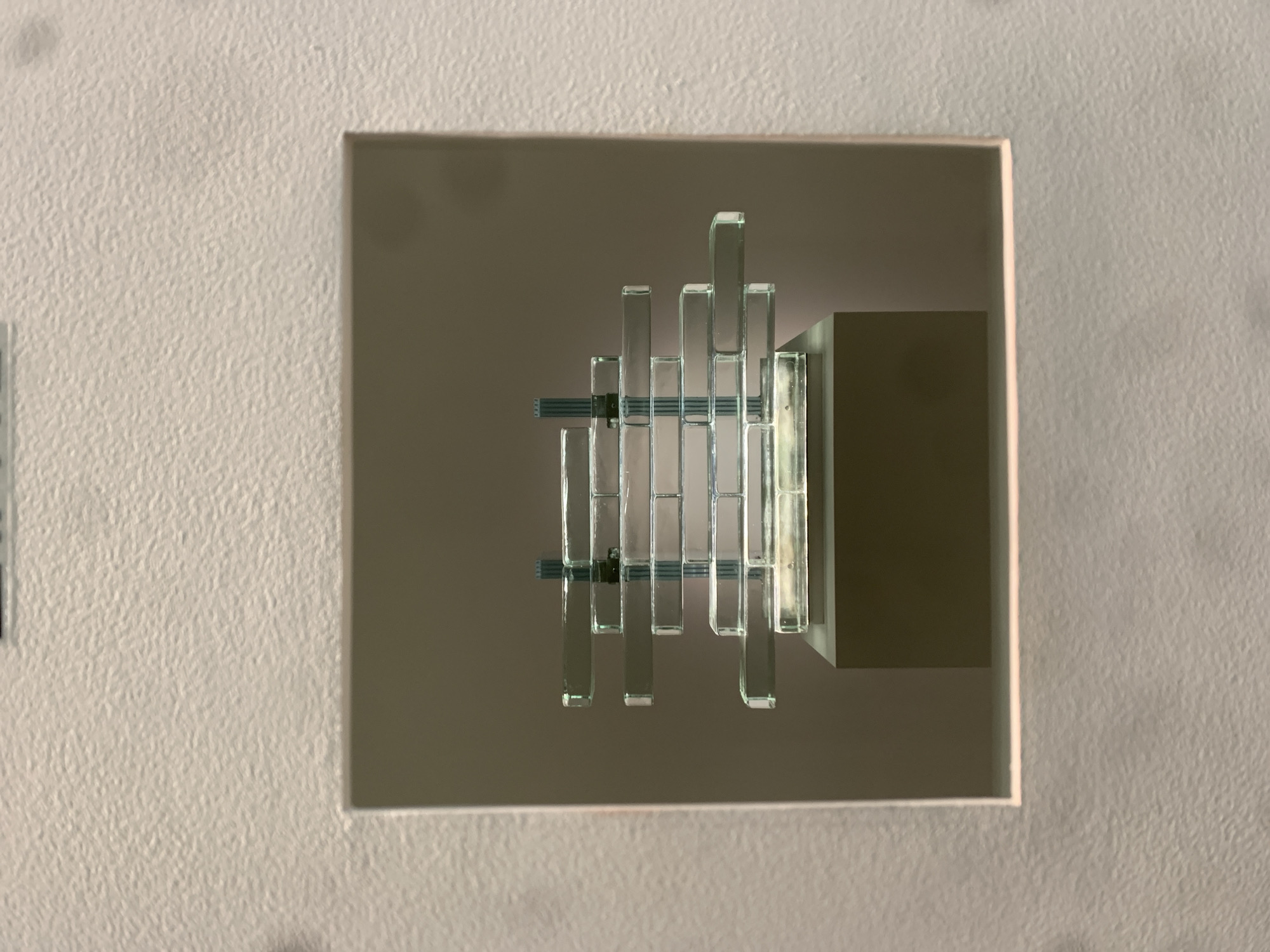 The image depicts a light fixture recessed into a textured, light-colored wall. The light fixture is square, with a series of vertical glass bars of varying heights arranged in a row. Two horizontal bars intersect the vertical pieces at the center. A tan or gray rectangular element appears to the right of the glass bars, presumably housing the electrical components. The light fixture appears to be illuminated, casting reflections on the glass. The surrounding wall has a rough, stucco-like texture. No people are visible in the image. The location is not discernable from the image, thus I cannot determine if it is in Moscow, Russia.