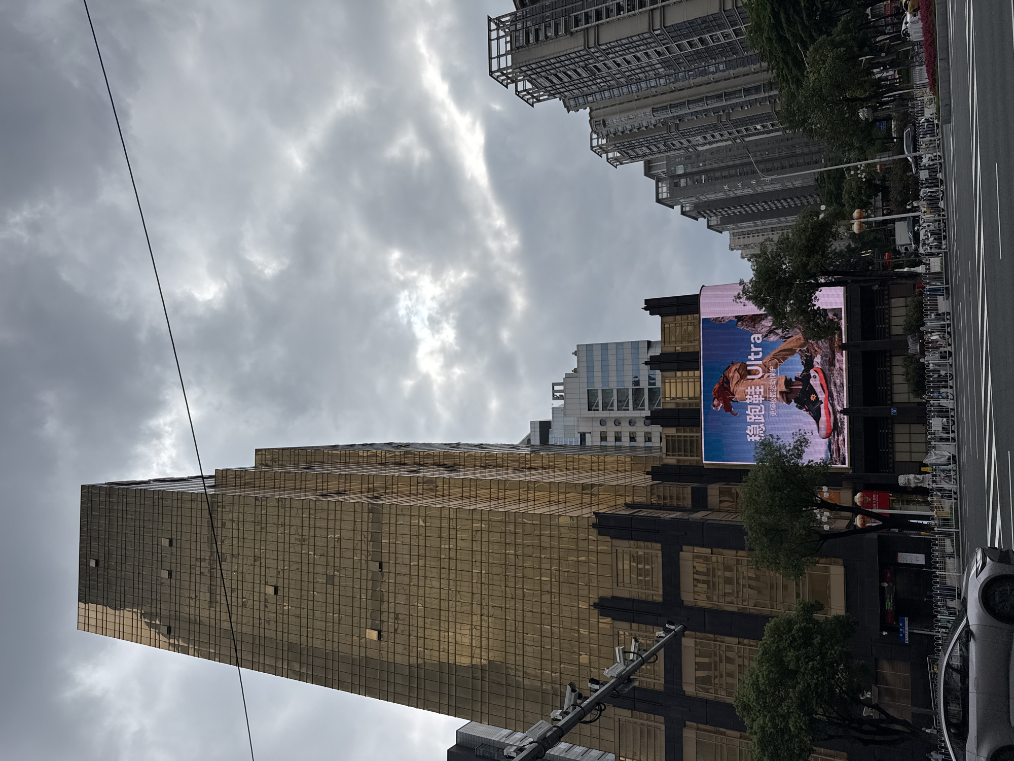 The image captures a wide street scene in Tianhe District, China, under a cloudy sky. A prominent golden skyscraper dominates the left side of the frame, its facade covered in glass tiles that reflect the overcast sky. To the right of the skyscraper, a large digital billboard displays an advertisement for "Ultra" running shoes, featuring a close-up of a shoe with red accents. Several other modern high-rise buildings are visible in the background. The street in the foreground is paved with asphalt and marked with white lane dividers. A silver car is parked on the left side of the street. A white fence runs along the sidewalk, behind which a row of parked electric scooters and bicycles are visible. Trees with green foliage are scattered throughout the scene, interspersed with streetlights and some decorative lion statues. People are present in the scene, walking on the sidewalk and possibly near the vehicles, but their features are not distinct. The overall impression is of a busy urban environment on a day with limited sunlight.