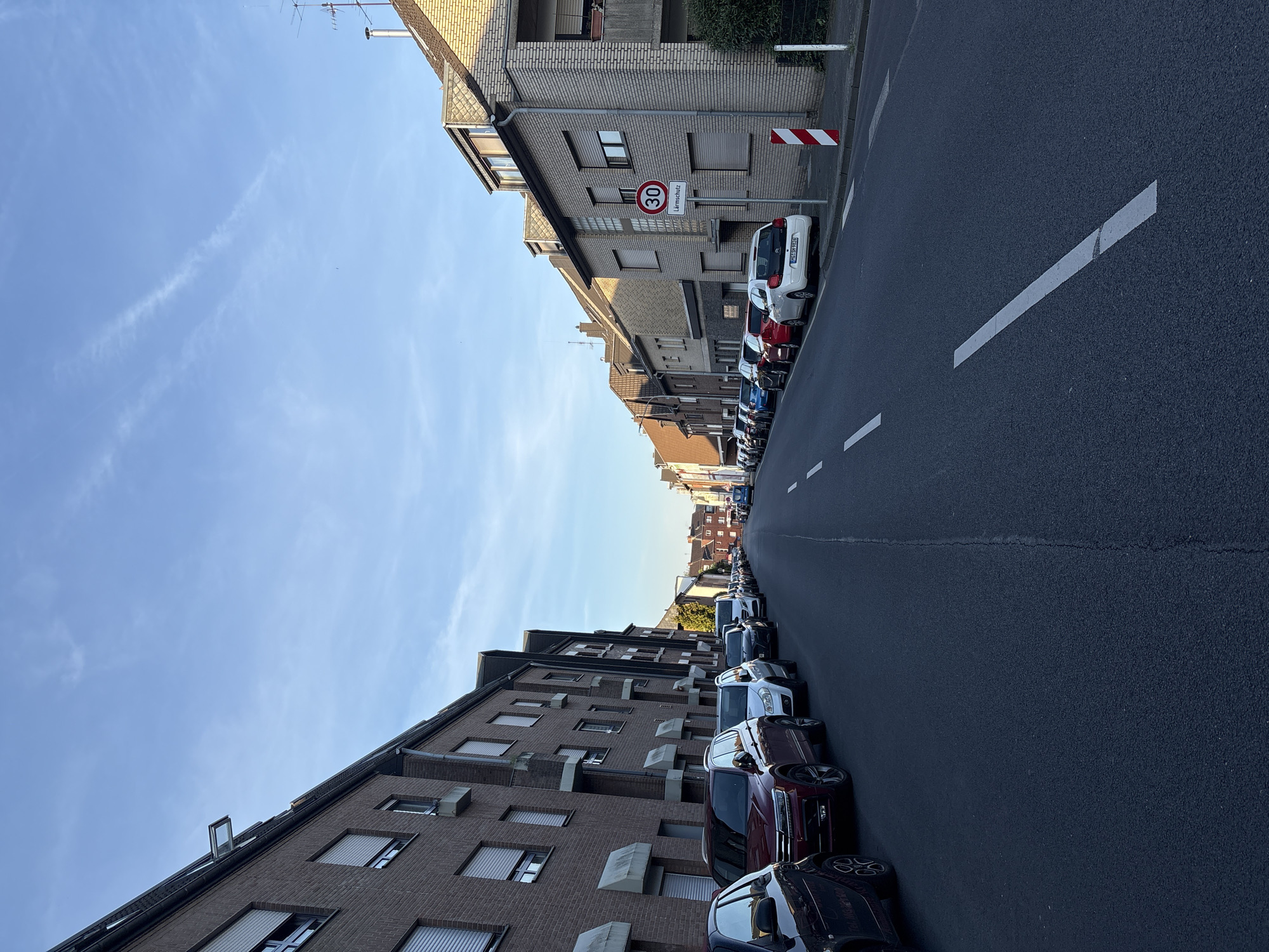 This image depicts a street in Mönchengladbach, Germany, characterized by parked cars lining both sides of the asphalt road. The buildings along the street are primarily brick apartment buildings, with variations in architectural style. A "30" speed limit sign is visible, indicating a 30 km/h speed restriction. White painted lines demarcate the traffic lanes. The sky is blue with scattered clouds. There are no people evident in the photograph.