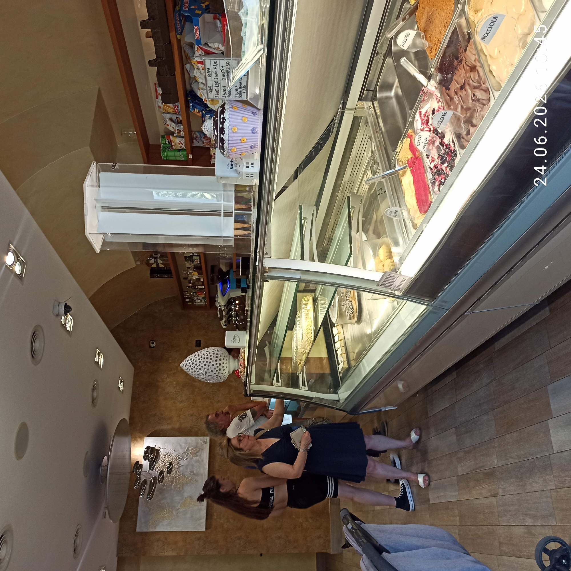 In Bitonto, Italy, a bright, modern ice cream parlor is depicted with an overhead perspective. The scene is dominated by a refrigerated display case filled with a vibrant assortment of gelato flavors, ranging from creamy vanilla and hazelnut to rich chocolate and fruit-infused options. Labels such as "Nocciola" (hazelnut), "Cioccolato" (chocolate), and "Biscotto" (biscuit) are visible.
In the foreground, three individuals are interacting with the gelato display. Two women and one man appear to be customers, with one woman holding a small bag and the other gesturing towards the flavors. A portion of a baby stroller is also visible, suggesting a family outing.
The walls of the parlor are a warm, earthy tone, with a textured finish. Decorative elements include a contemporary map art piece on the wall and a sculptural light fixture on the ceiling. The floor is made of wooden planks.
The lighting is bright and artificial, likely from the display case and overhead lights, suggesting an indoor setting, possibly during the day or early evening. The timestamp "24.06.2023 10:45" is superimposed on the image, indicating the time the photograph was taken. The overall atmosphere appears casual and relaxed, typical of a visit to an ice cream shop.