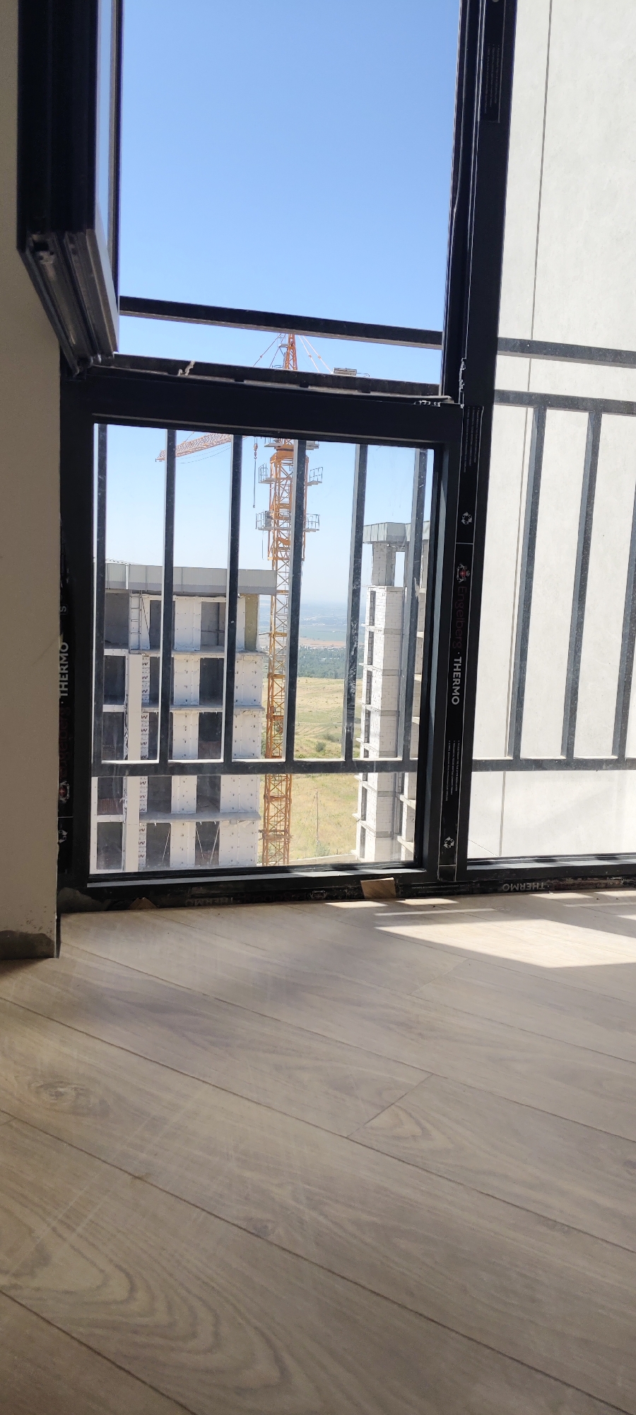 This is a vertical, eye-level, medium shot photograph taken from inside a building, looking out through a large, open window. The perspective is from a low angle, with the floor in the foreground and the sky in the background.
The foreground consists of a light brown, wood-planked floor with visible grain patterns and some light scuff marks. The floorboards are laid horizontally relative to the frame.
The midground and background are dominated by a view of a construction site. A large, orange construction crane is prominent, with its lattice structure clearly visible against the bright blue sky. To the left of the crane, the unfinished facade of a building is visible, with concrete pillars and white concrete blocks forming its structure. The building appears to be multi-story and is in the early stages of construction. Further in the distance, a hazy, green landscape is visible, suggesting a suburban or rural area outside of a city.
The window through which the scene is viewed has a dark gray frame. The lower part of the window appears to be a sliding or openable section, with a black frame and vertical bars extending from the top to the bottom of the window pane. On the vertical frame, there are some labels, one of which appears to read "THERMO."
The lighting suggests it is daytime, with bright sunlight casting shadows and illuminating the scene. The sky is clear and blue with no clouds. The overall impression is a snapshot of urban development in progress.
While the prompt asks for the location to be Aktash, Uzbekistan, there are no visual cues within the image itself that definitively confirm this specific location. The scene depicts a generic construction site that could be found in many urban or developing areas.