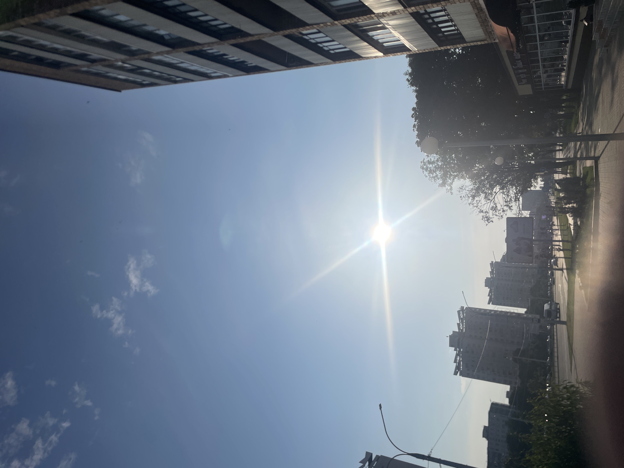 The image depicts a street scene in a city under a bright, sunlit sky with scattered clouds. On the right, a tall, multi-story brick building with numerous windows dominates the foreground. To the left and in the mid-ground, several similar apartment buildings are visible, as well as trees and streetlights. A road with a few vehicles is also present. The sun is positioned in the upper center of the frame, creating a strong glare and lens flare effect. The environment suggests it is daytime in an urban setting.