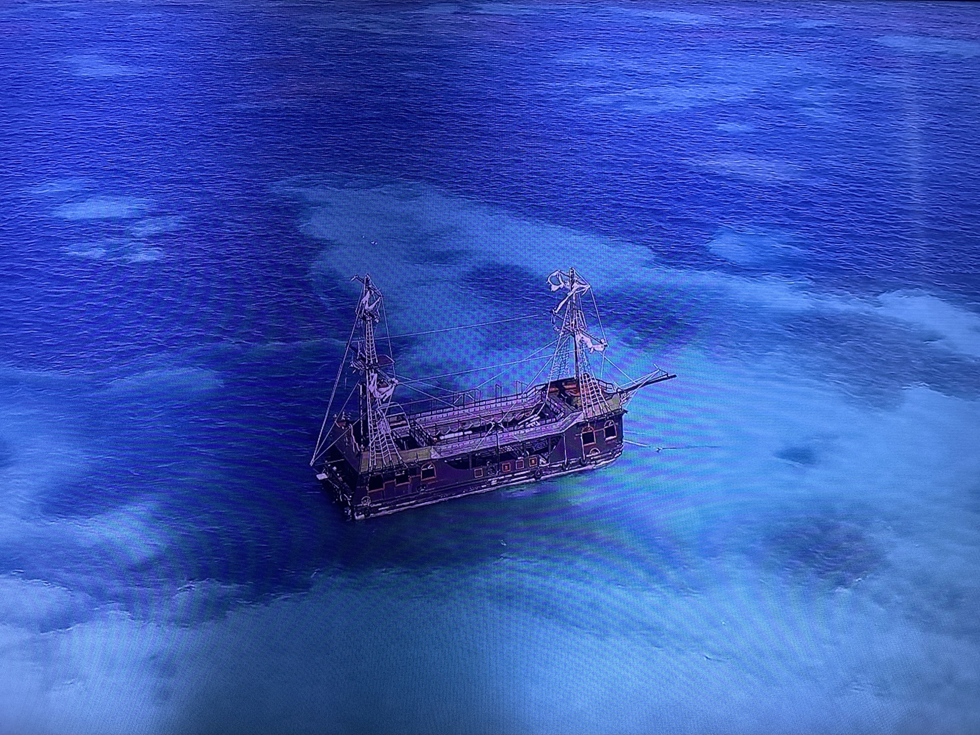 This media file shows a digital rendering of a pirate ship sailing in clear, blue tropical waters. The ship is a large, multi-masted vessel with a dark hull, several decks, and intricate rigging. Its sails are furled, suggesting it is either at anchor or moving slowly. The water is shallow enough in some areas to reveal lighter sand or coral formations beneath the surface, indicating a reef or shoal. The overall scene evokes a sense of adventure and the allure of the open sea, typical of pirate lore. The lighting suggests it could be daytime, possibly late morning or early afternoon, with the sun high enough to illuminate the underwater features. There is no text visible in the image, nor any explicit indication of the time of day or weather conditions beyond the clarity of the water and the sunlight. The scene is set in an unknown tropical location.