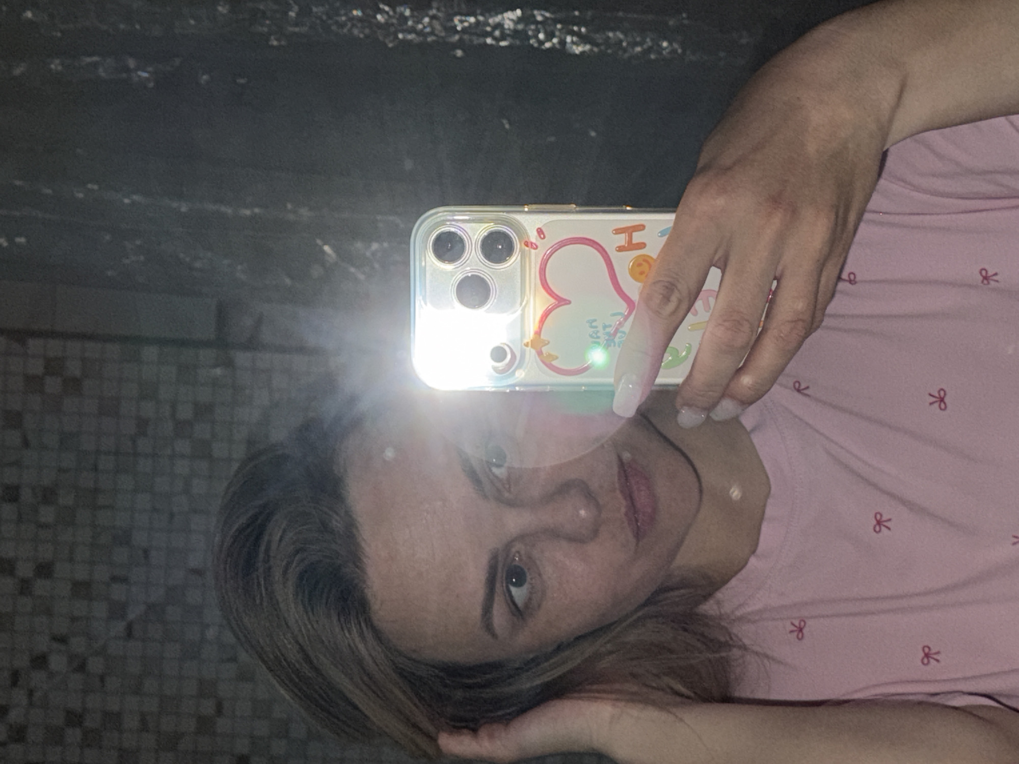 A woman is taking a selfie in front of a bathroom mirror. The mirror reflects a tiled wall and a section of the ceiling. She is holding an iPhone with a decorative case. The woman has fair skin, shoulder-length blonde hair, and is wearing a pink shirt with small bow patterns. The location of the image is unknown.