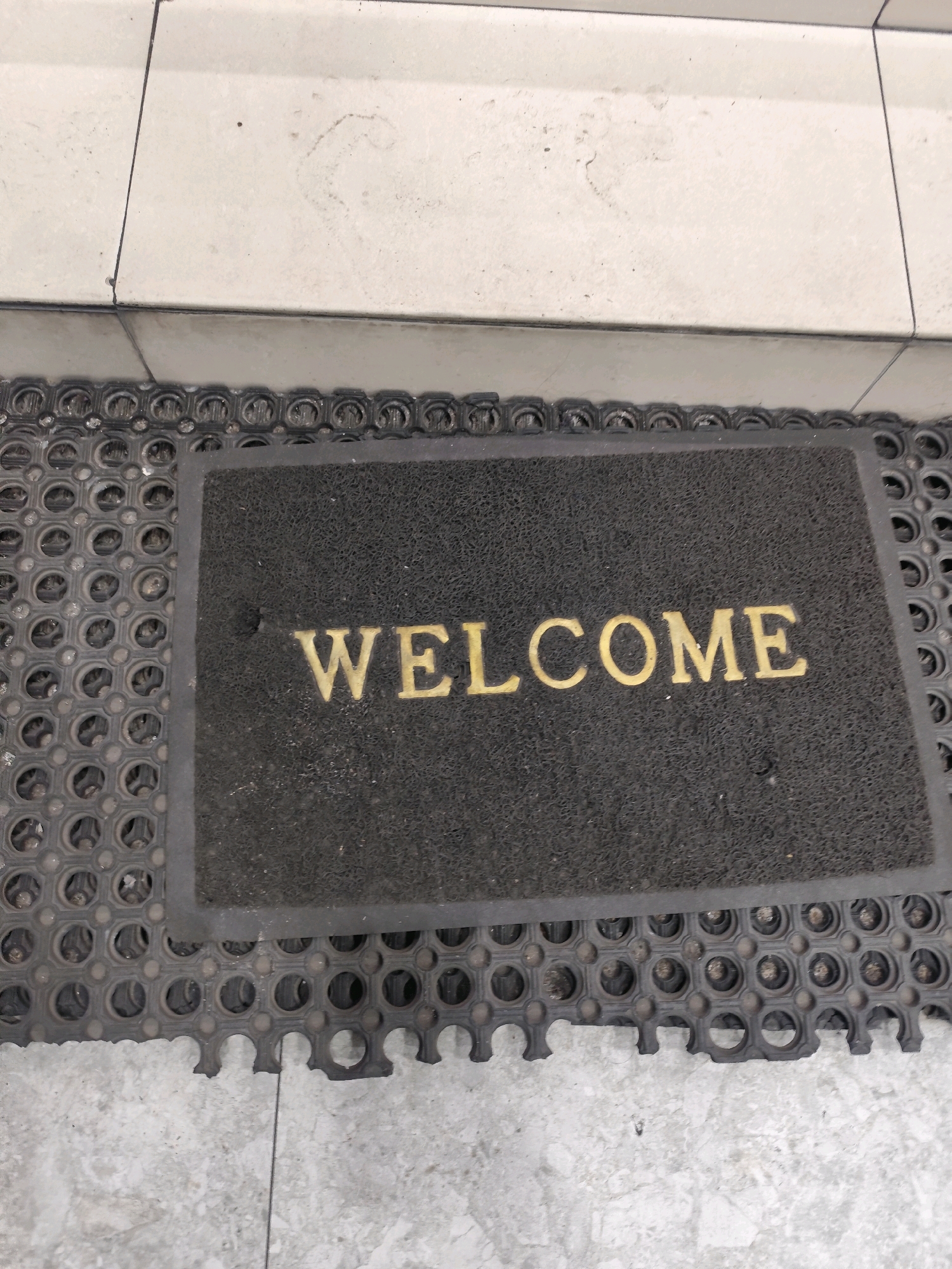 A black doormat with the word "WELCOME" embossed in gold letters is placed on a grey textured mat. The mats are situated on a tiled floor. The scene is a close-up, low-angle shot. There are no people or animals visible. The environment appears to be an interior or exterior entryway. The location is the city of Palangka Raya, Indonesia.
