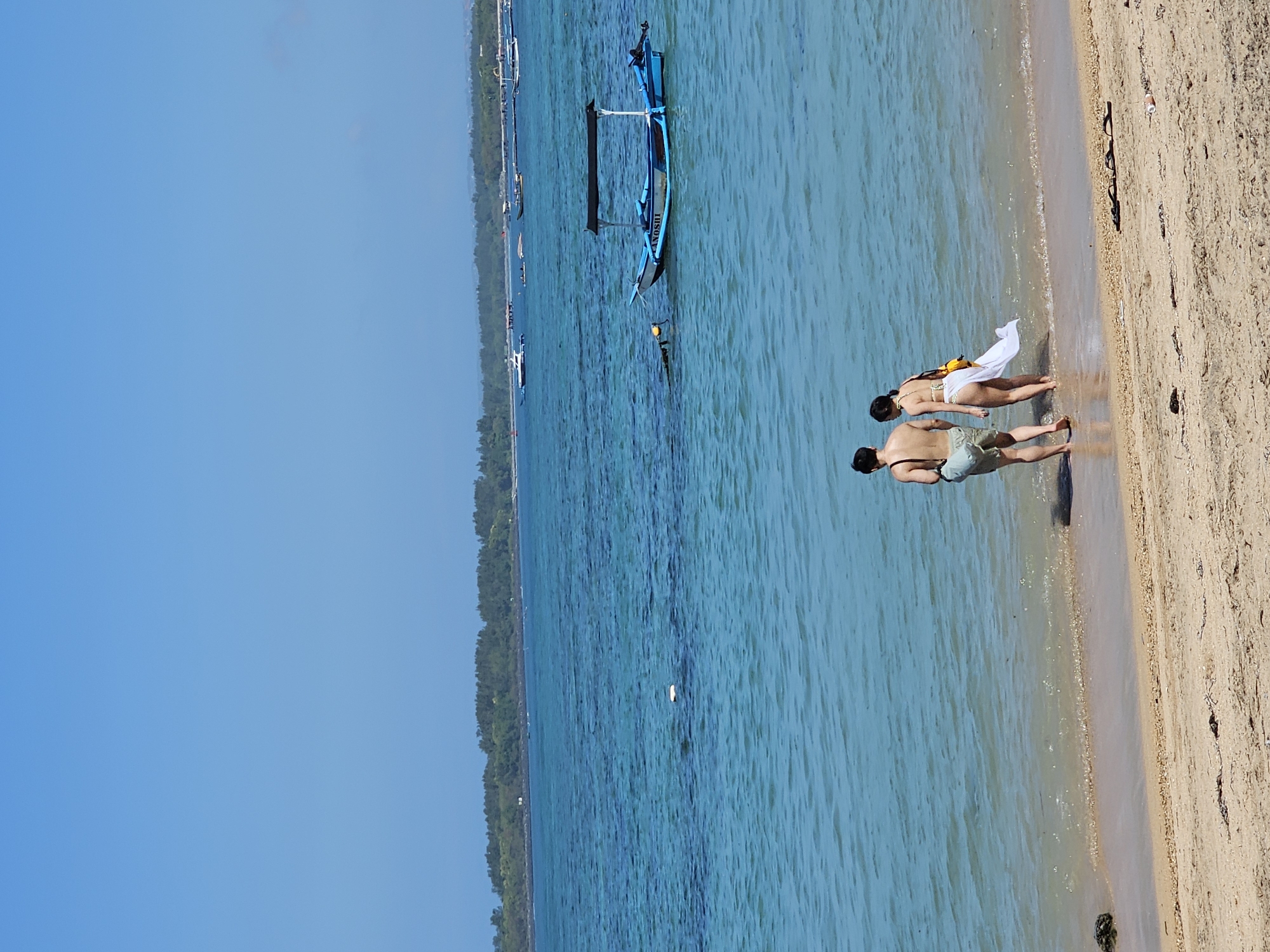 A couple walks into the clear turquoise water on a sandy beach in Denpasar, Indonesia. The man is wearing light-colored shorts and carrying a bag over his shoulder, while the woman is wearing a white bikini and a sheer white sarong draped over her shoulders. The water is shallow near the shore, with gentle waves lapping at their feet. In the background, a traditional Balinese outrigger boat with a canopy is anchored in the sea, along with other boats in the distance. A lush green treeline borders the horizon, suggesting a tropical landscape. The sky is a clear, bright blue, indicating a sunny day. The beach itself is a pale golden sand, with some scattered debris.