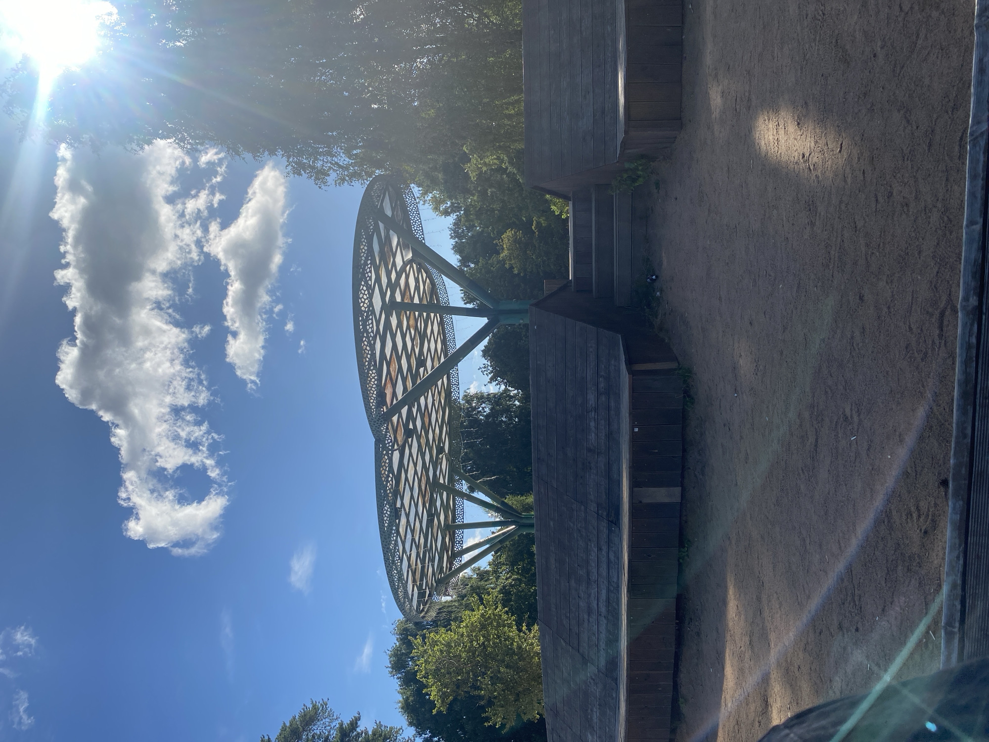 The image shows an outdoor amphitheater or stage area with tiered wooden seating in a park. A distinctive, modern canopy with a patterned roof supported by tree-like structures stands behind the stage. The ground in front of the seating is covered in fine gravel or sand. Lush green trees surround the area, and a bright, sunny sky with scattered clouds is visible above. The scene appears to be located in Saint Petersburg, Russia. The lighting suggests it is daytime, possibly late afternoon due to the angle of the sun and the lens flare. There are no people visible in the frame.