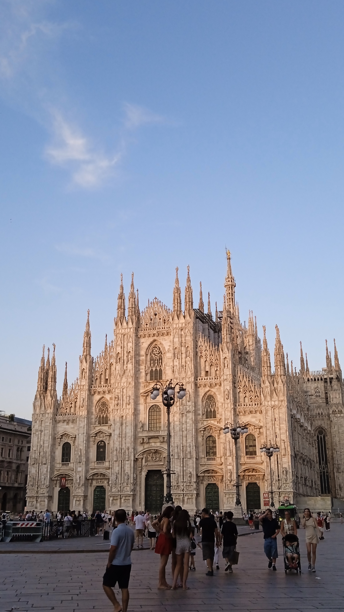 The image shows the Duomo di Milano, a cathedral located in Milan, Italy. The cathedral is a large, ornate Gothic building with numerous spires and statues. In the foreground, there is a plaza with several people walking around. The sky is clear and blue. The lighting suggests it is late afternoon or early evening. The people are casually dressed and appear to be tourists.