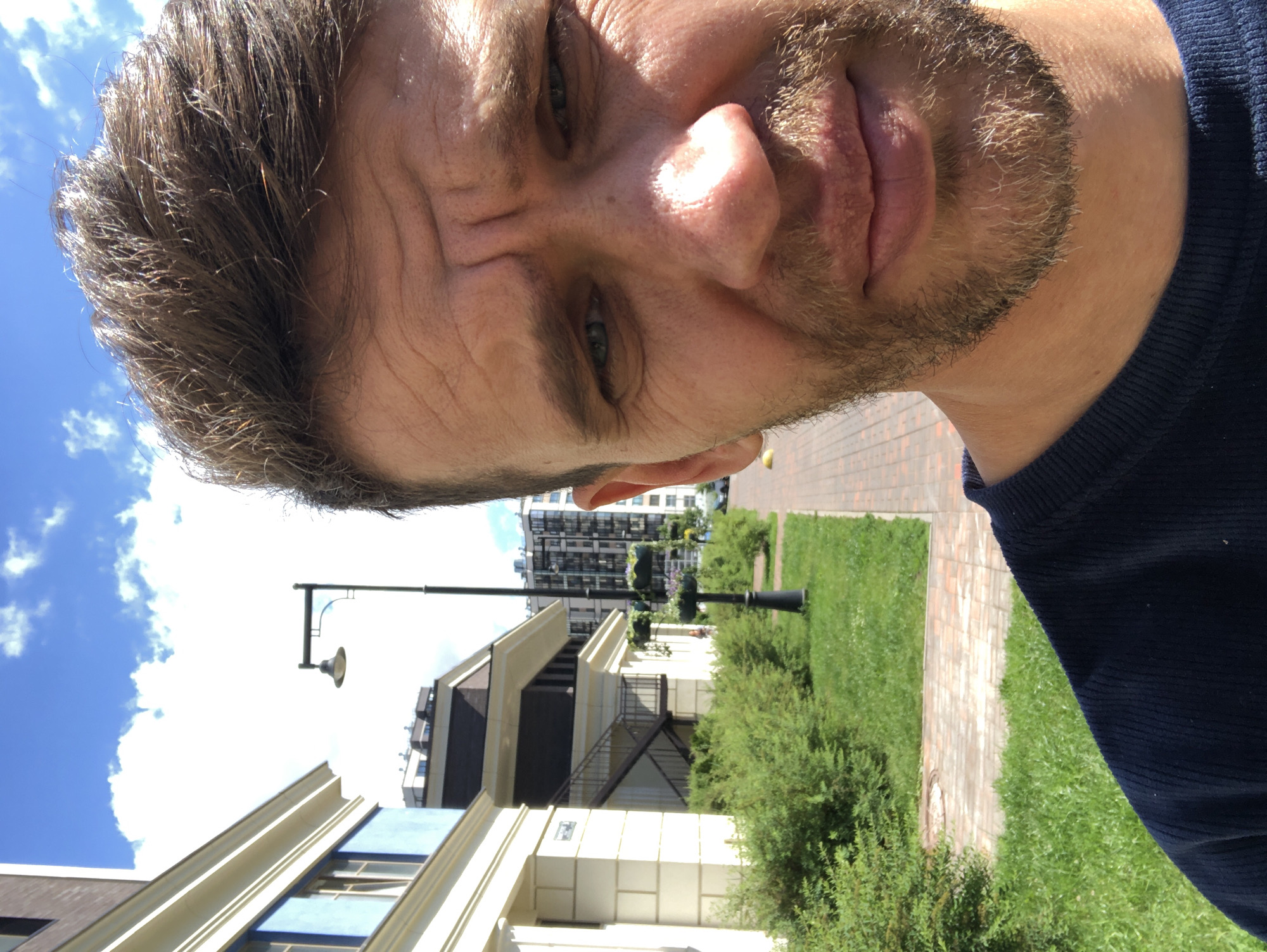 A selfie is taken outdoors on a sunny day in a residential area. The subject, a man with light brown hair and a beard, is positioned in the right half of the frame, looking directly at the camera. His expression is neutral. In the background, to the left, is a modern apartment building with light-colored walls and blue accents. Adjacent to the building is a paved walkway lined with manicured green bushes and grassy areas. A street lamp with a planter box hangs above a low bush in the middle ground. Further in the background, other multi-story buildings are visible, including a darker, modern apartment complex. The sky above is a bright blue with scattered white clouds. The location is identified as Saint Petersburg, Russia.