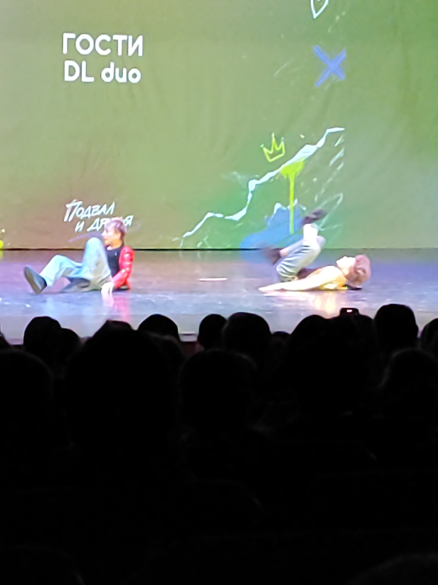 A stage performance in Fryazino, Russia is captured in this image. Two individuals are performing on stage, with the backdrop featuring the words "ГОСТИ DL duo" and other graphics. One performer is seated while the other is lying down, with their legs in the air. A crowd of people is visible in the foreground, watching the performance. A mobile phone is being used to record the performance.