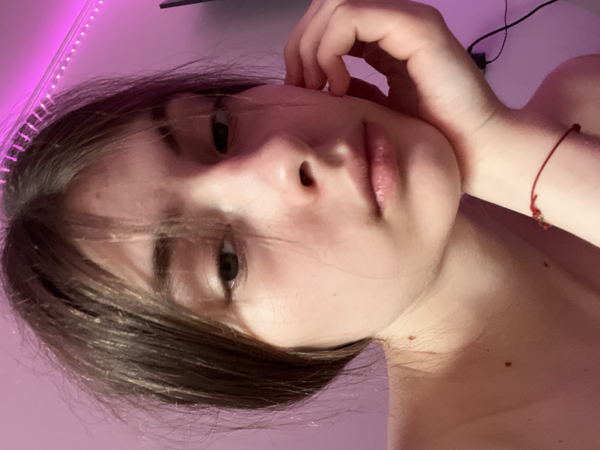 A close-up, slightly angled, portrait of a fair-skinned person with dark brown hair. The person's face is the main subject, with their right hand near their face. A thin red bracelet is visible on their right wrist. The background is a blurry pink, suggesting an indoor setting with pink lighting, possibly a strip of LED lights. A black cable is partially visible in the upper right corner. The location in Saint Petersburg, Russia, cannot be determined from the given image.