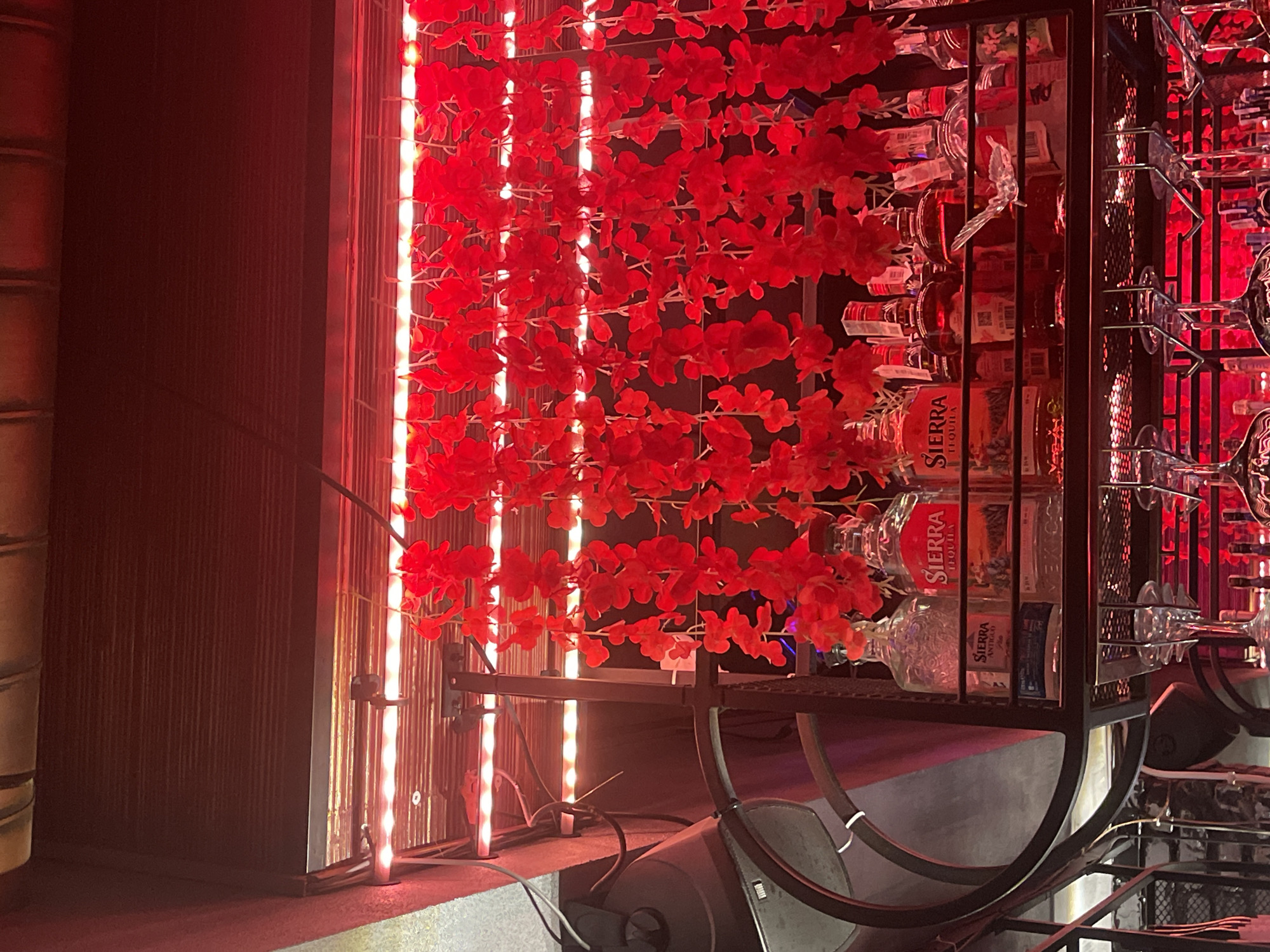 The image shows a shelf in a dimly lit bar, illuminated by red LED lights. The shelf holds several bottles of alcohol, including at least two bottles of Sierra Tequila. In front of the bottles, there are hanging strings of red artificial flowers. Below the shelf, wine glasses are inverted and hung on racks. The background is dark, with some metallic structures visible. No people are present in the image. This image does not provide any information about the city of Poltava, Ukraine.