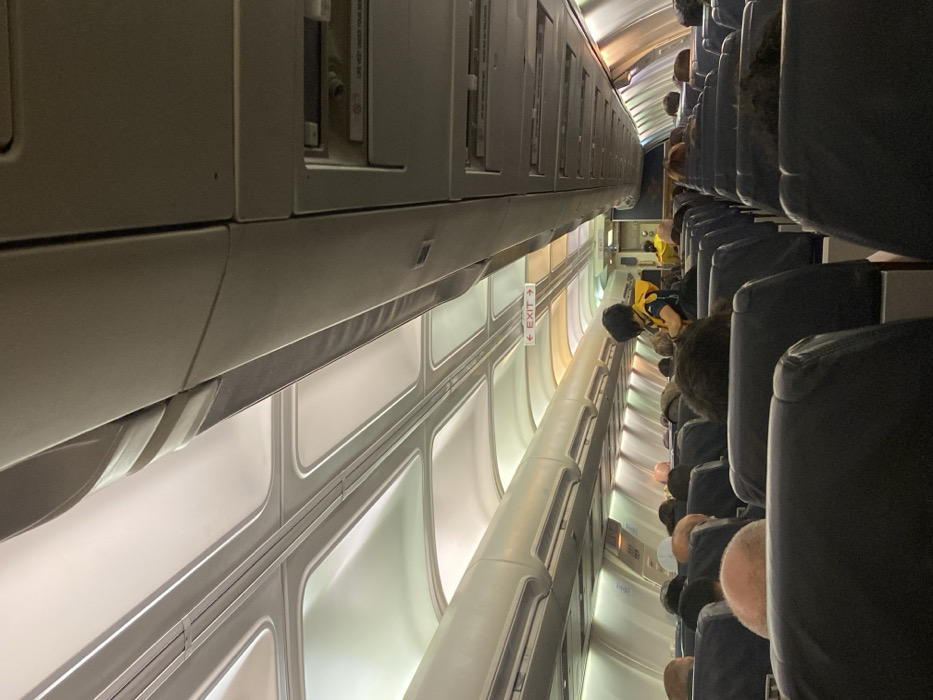 The scene is the interior cabin of a commercial aircraft, presumably located at Athens International Airport "Eleftherios Venizelos" (ATH) within the Paiania Municipal Unit, Greece. The perspective is from a passenger seat, looking forward down the central aisle. Numerous passengers, primarily visible from the back of their heads and shoulders, occupy rows of dark blue/grey seats on both sides of the aisle. Approximately midway down the cabin, a person with dark hair, wearing a black top and an orange/yellow life vest, stands facing away from the camera, likely a flight attendant conducting a safety demonstration. Further forward in the aisle, another individual in a yellow garment is partially visible near the aircraft's front or galley area. The cabin ceiling features illuminated panels and overhead compartments, extending the length of the visible area. A red-on-white "EXIT" sign with a left-pointing arrow is illuminated above the aisle in the distance. Passenger service units are mounted below the overhead compartments.