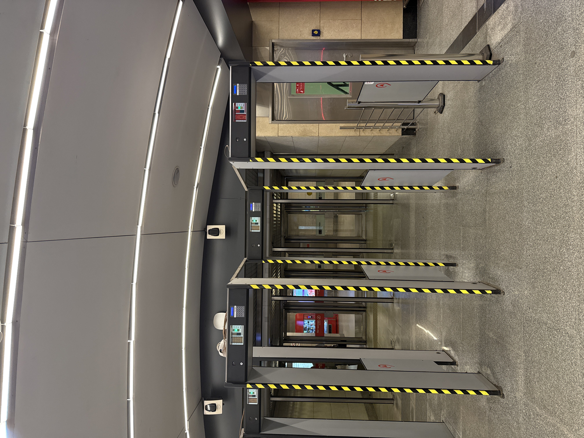 The image shows a row of turnstiles inside a subway station. The station appears to be located in Moscow, Russia, based on the Cyrillic text on a vending machine visible through the turnstiles. The turnstiles are gray and feature yellow and black striped safety markings. Each turnstile has a control panel with buttons and a display screen. A person wearing a white hat and ear protection is visible near one of the turnstiles. The environment includes a gray floor and a curved ceiling with recessed lighting. Beyond the turnstiles, there are doors and a vending machine.