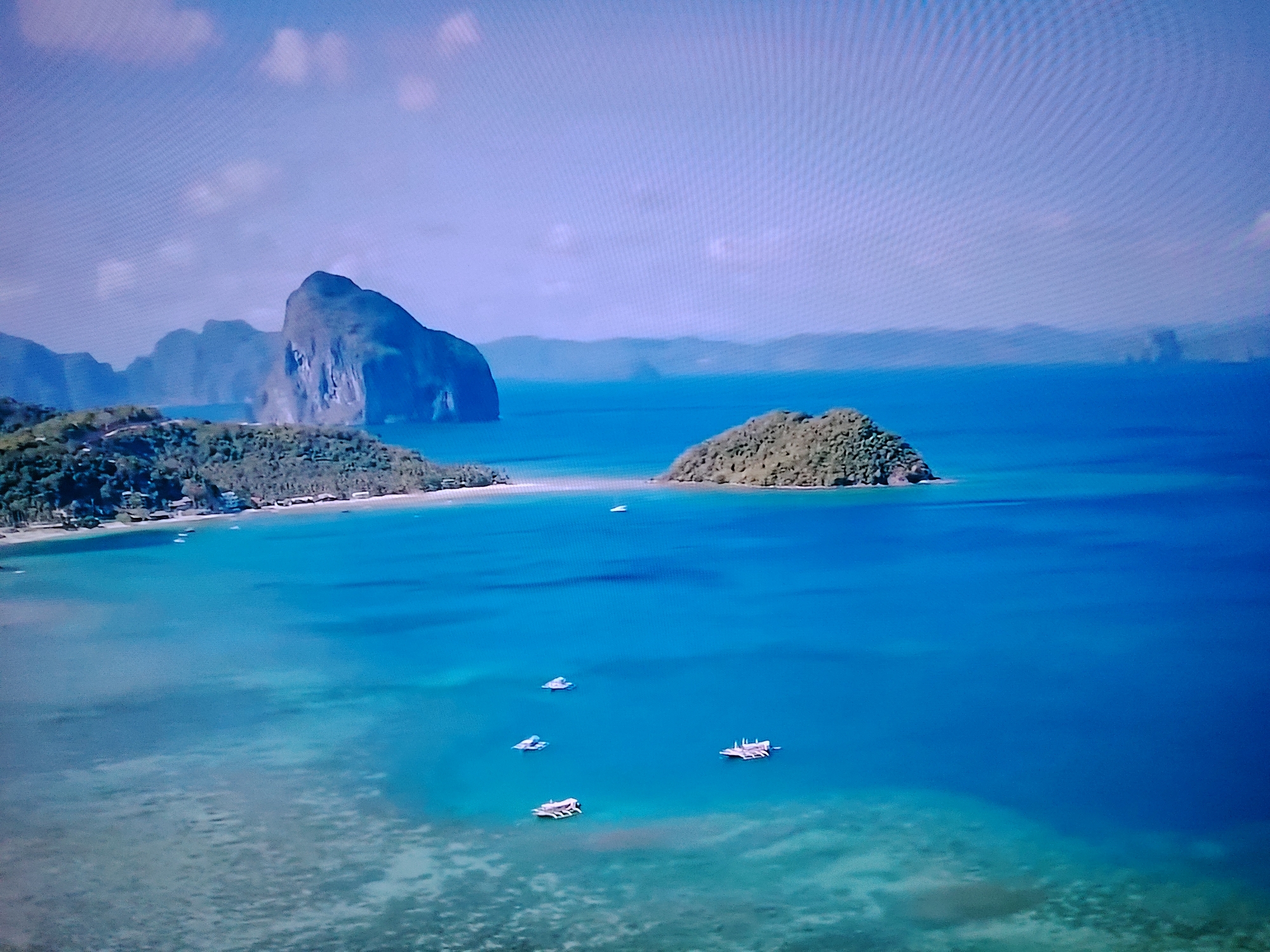 The image is an aerial view of a tropical coastline with several islands and boats. The water is a vibrant blue, transitioning to lighter turquoise near the shore, revealing submerged coral reefs. In the foreground and mid-ground, several small boats are scattered across the water. In the mid-ground to the left, a long, narrow island with lush green vegetation and a sandy beach is visible, with some buildings nestled amongst the trees. To the left of this island, a large, imposing rock formation with steep cliffs rises dramatically from the sea. In the background, distant, hazy islands form a layered horizon. The sky is a pale blue with scattered white clouds. The scene depicts a serene and picturesque coastal landscape.
There is no indication in the image that this location is in Huntingdonshire, United Kingdom. The landscape is characteristic of a tropical or subtropical region.