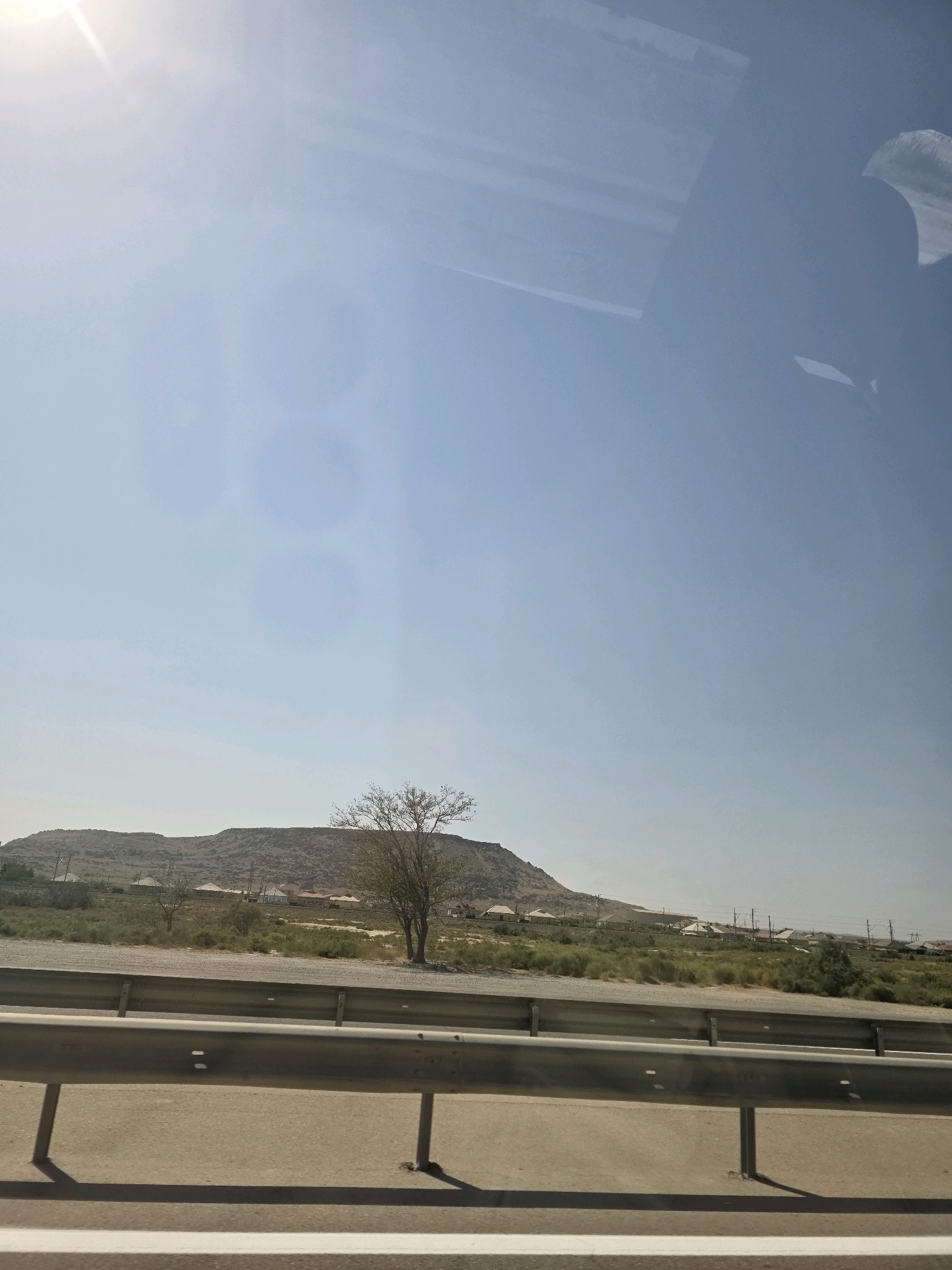 A medium shot captures a scenic view of the outskirts of Ghobustan, Azerbaijan, bathed in the bright light of a sunny day. The image is dominated by a vast expanse of clear blue sky, with the sun a prominent, lens-flare-inducing presence in the upper left quadrant. A hint of a reflection, possibly from a window, is visible in the upper right corner, obscuring a portion of the sky.
The mid-ground of the image features a low, undulating hillside covered in sparse vegetation, appearing dry and somewhat barren. A solitary, bare-branched tree stands on the crest of the hill, its silhouette stark against the bright sky. Below the hill, a cluster of residential buildings, characterized by their light-colored roofs, forms a small settlement. The houses appear to be single or two-story structures, spaced out amidst arid greenery.
In the foreground, a metallic guardrail runs horizontally across the image, separating a paved road from the undeveloped land. The guardrail is gray and shows signs of weathering. The road surface is a light brown or tan color, with a white line marking its edge. Shadows from the guardrail posts are cast on the road, indicating the direction of the sunlight. The overall environment suggests a semi-arid, rural landscape. There are no people or animals visible in the frame.