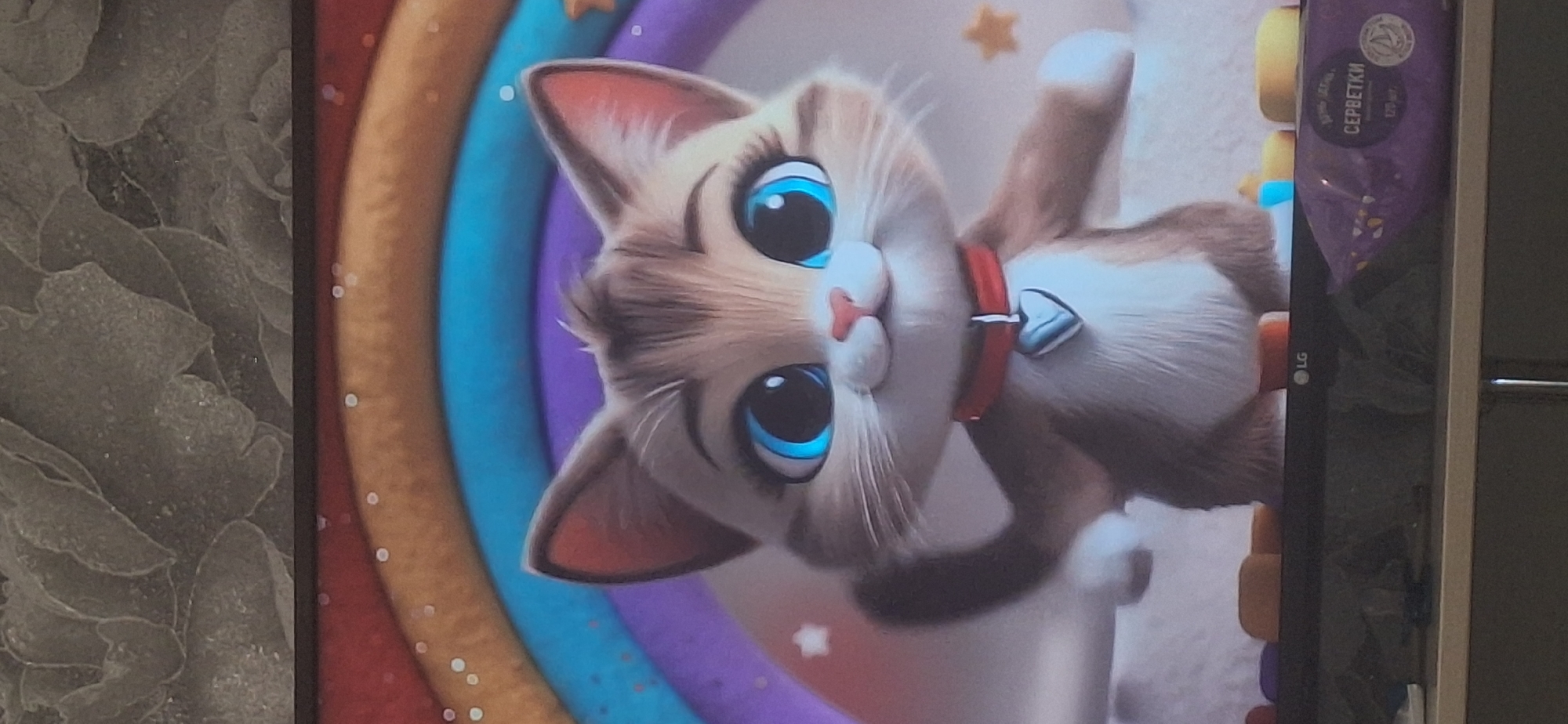 The image displays a digital rendering of a cartoon kitten with large blue eyes, a pink nose, and a red collar with a heart-shaped pendant. The kitten is depicted in front of a rainbow with multiple colored arcs and sparkling stars. The background shows a textured surface, possibly wallpaper, at the top. Below the kitten, the image captures the top of a television screen displaying the cartoon, with some objects on the surface in front of it, including what appears to be a package of wet wipes. The scene is indoors, and the overall impression is that of a child's room or entertainment area. There is no visible indication of the city of Bila Tserkva, Ukraine, nor any specific geographical markers.