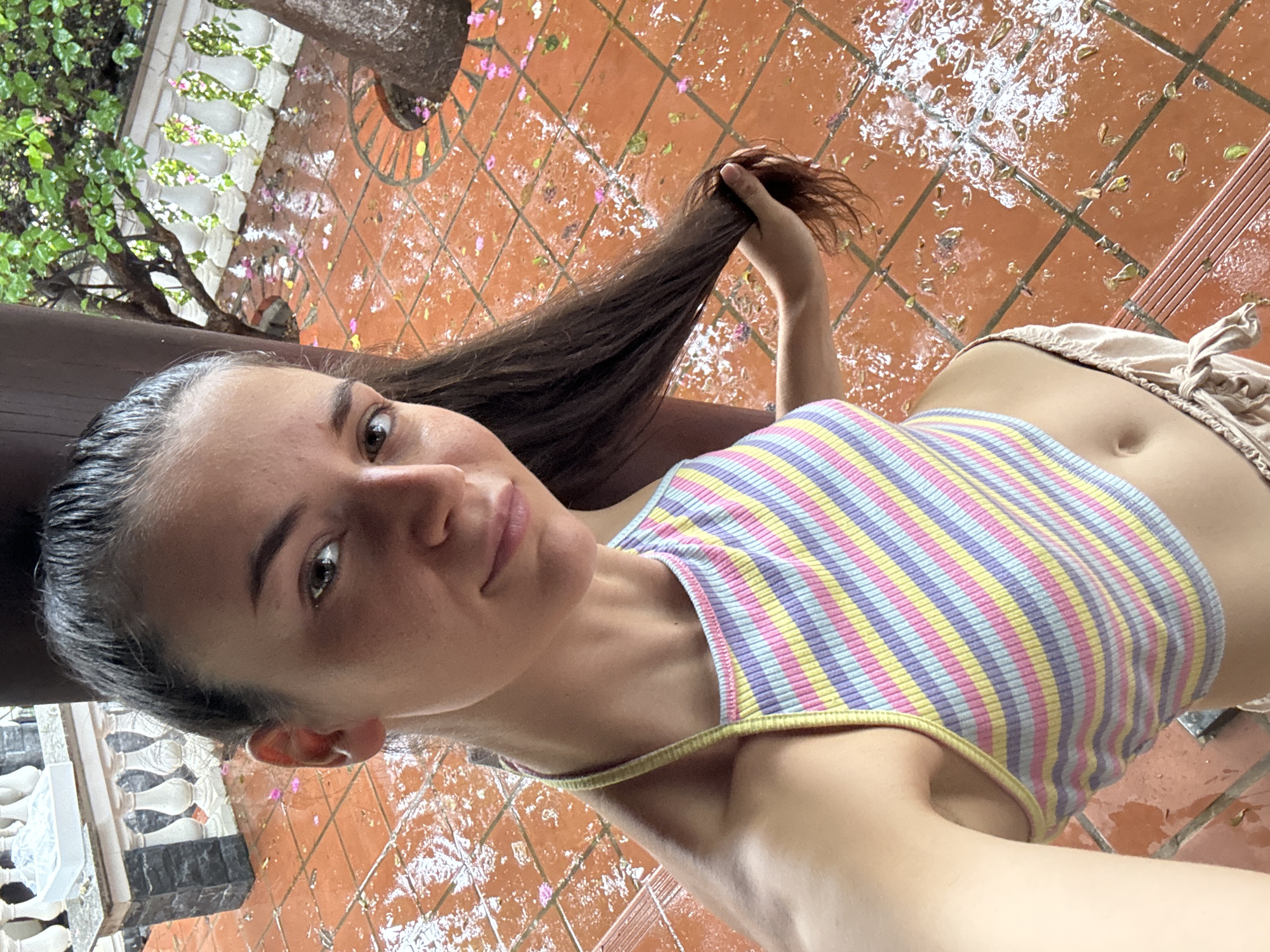 A woman is taking a selfie outdoors on a wet, tiled patio. She is holding a section of her long, brown hair. She is wearing a striped tank top and tan shorts. The patio appears to be in a courtyard area with plants and trees visible. This may be in the city of Mũi Né, Vietnam.