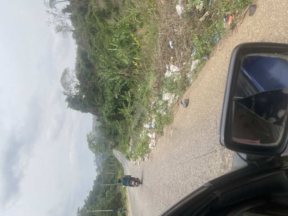 The scene depicts a roadside environment in Oka-Akoko, Nigeria, viewed from inside a vehicle. A paved asphalt road, showing minor cracks and wear, extends into the distance, appearing to follow an undulating terrain.

In the midground, a single individual rides a dark-colored motorcycle away from the viewer on the left side of the road. The rider is wearing dark clothing. To the right of the asphalt road, a distinct and extensive accumulation of refuse is present along the dirt and grass shoulder. This refuse consists primarily of white and transparent plastic bags, discarded packaging, and other assorted litter, scattered amidst weeds and dry vegetation. A single dark plastic bag is positioned on the paved shoulder closer to the foreground.

Beyond the littered shoulder, a dense embankment or hillside covered in tropical vegetation, including broad-leafed plants and various trees, rises significantly. Hilly terrain with similar vegetation is visible in the background. On the left side of the road, several streetlights with blue and white poles are visible, extending into the distance. The sky overhead is overcast, with diffuse light. A portion of a blue vehicle side mirror, exhibiting a distinct crack, is visible in the lower right foreground, partially reflecting the interior of the viewing vehicle.