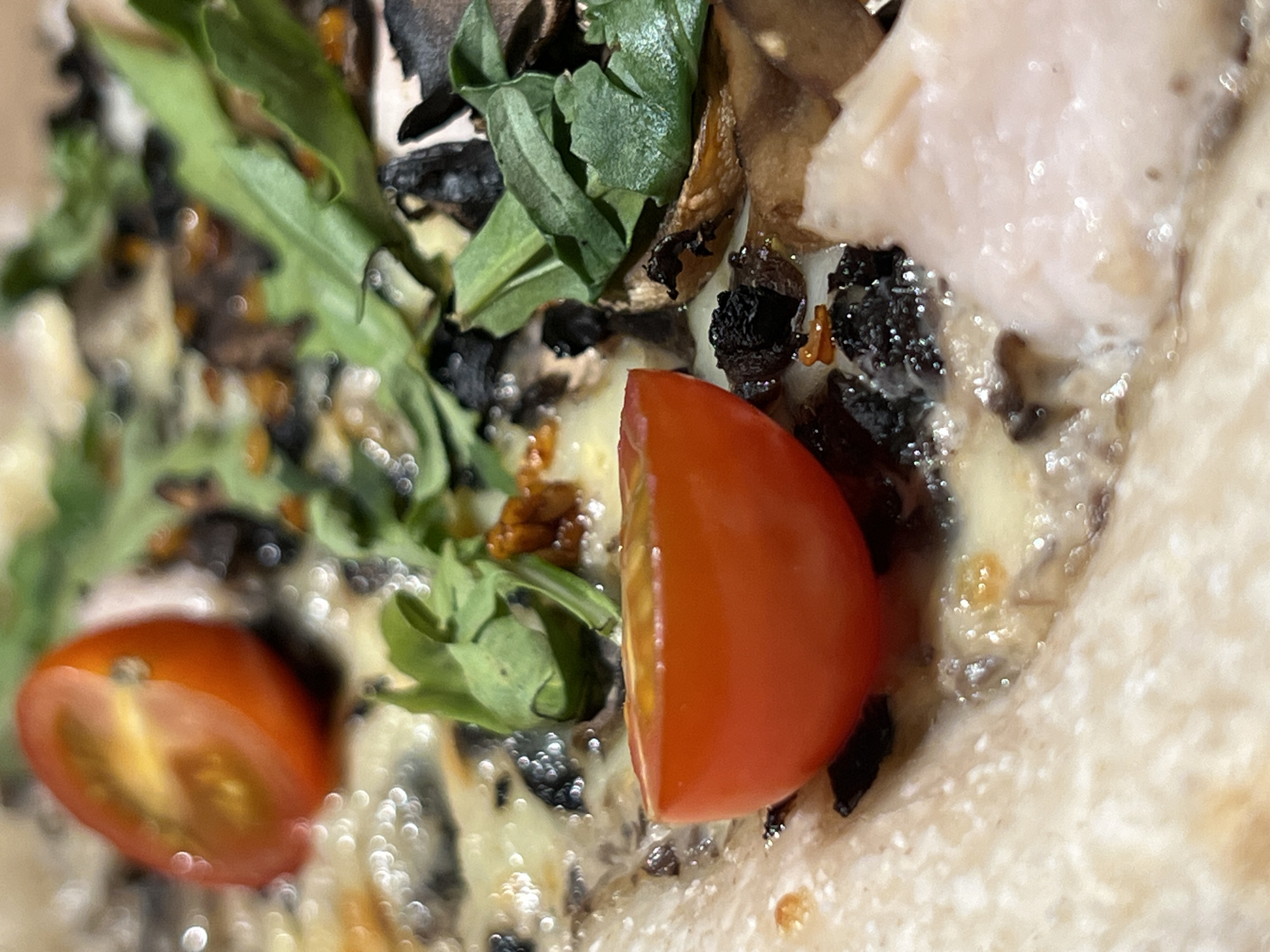 The image shows a close-up, overhead view of a pizza. The pizza has a thin crust and is topped with melted cheese, cherry tomatoes, mushrooms, and arugula. One cherry tomato is cut in half, revealing its seeds and pulp. Another half cherry tomato is visible in the background. Some pieces of meat, possibly chicken or ham, are also present on the pizza. The lighting creates a glossy effect on the cheese and vegetables, with bokeh highlights visible throughout the image. The specific location in Chaikovsky, Russia, is not discernible from the image content.