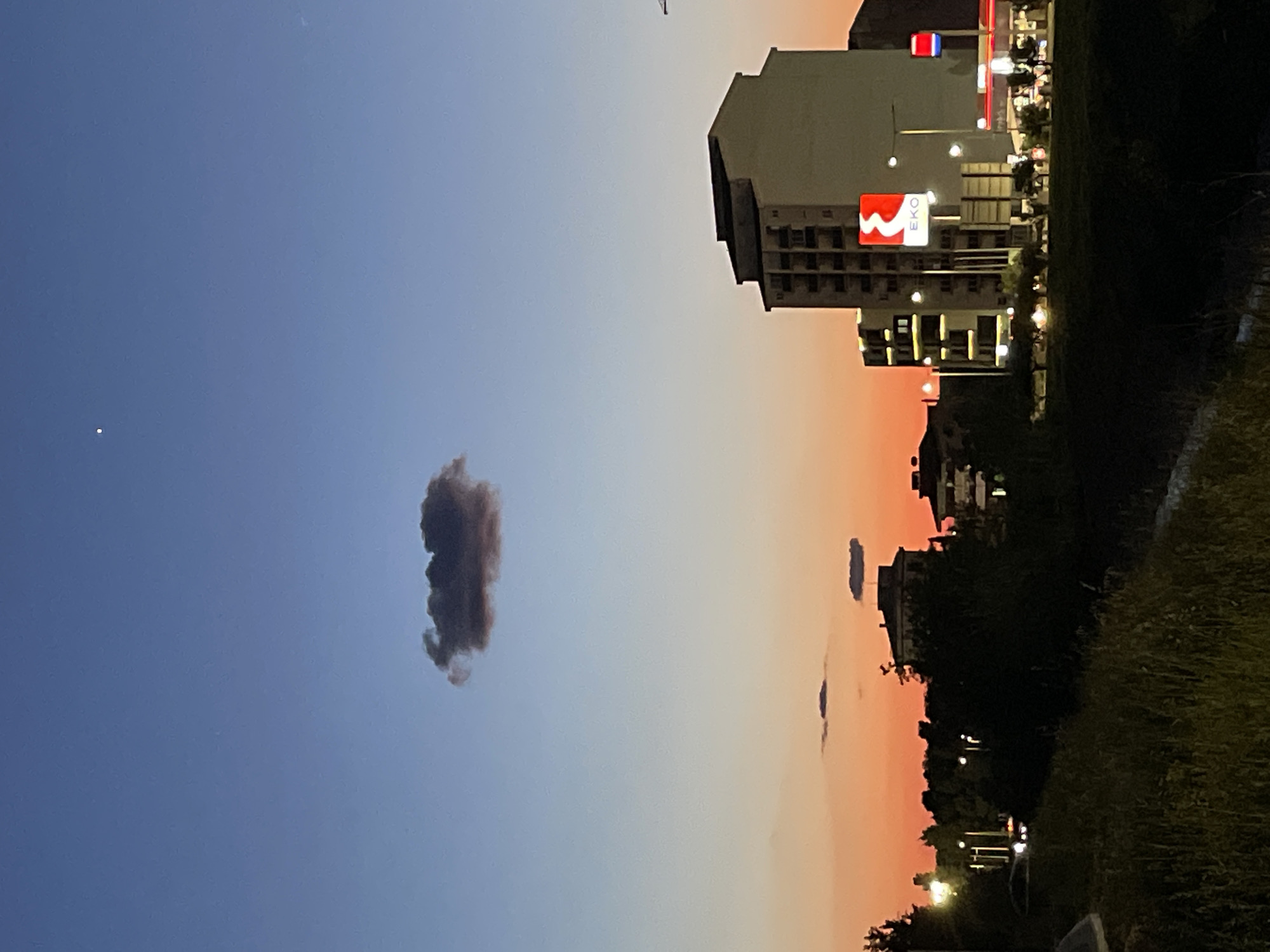 The image captures a dusk or twilight scene in an urban or suburban setting, likely in Bar, Montenegro. The sky dominates the upper portion, displaying a striking gradient from deep blue on the left, transitioning through lighter blue, then pale orange, to a rich reddish-orange hue on the right, suggesting either sunset or post-sunset glow.
In the upper left, a small, bright white dot, possibly a star or planet, is visible against the blue sky. A single, prominent dark grey cloud with an irregular, somewhat elongated shape floats in the mid-left sky. Further to the right, against the orange sky, several smaller, darker, and flatter cloud formations are visible.
Along the right side of the frame, several buildings are present. The most distinct is a multi-story, light-colored building with dark windows, featuring a prominent red square logo with white text that clearly reads "EKO" and a white wavy line above it. This building, along with others, shows illuminated windows and internal lights. Streetlights are also visible, casting glows on the foreground and illuminating parts of the structures.
The bottom of the image, particularly on the right, is filled with dark, silhouetted vegetation, including trees, bushes, and grass, indicating a roadside or park area. The artificial lights from the buildings and streetlights contrast with the dimming natural light. The overall mood is calm and serene, characteristic of the transition from day to night.
