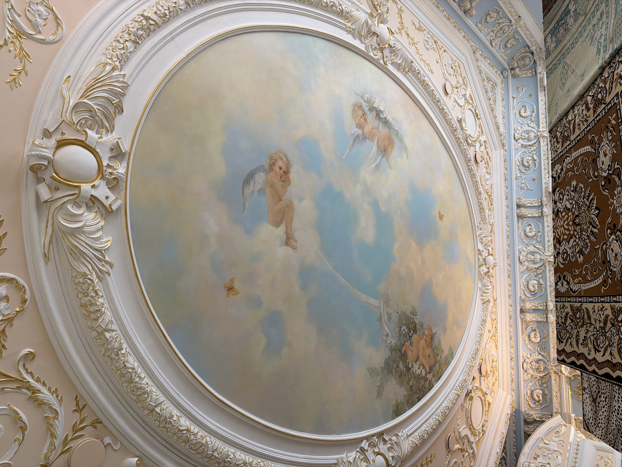 A detailed, ornate ceiling mural adorns a room, displaying a classical scene reminiscent of Renaissance art. The mural is oval and framed with layered, elaborately carved white and gold decorative elements. It depicts cherubic figures amidst painted clouds, evoking a heavenly or ethereal atmosphere. Two winged cherubs are prominent: one is suspended mid-air, while the other appears to be holding a garland of flowers. Small butterflies are also visible within the cloudscape.
The walls surrounding the mural feature similar ornate decorations, including relief patterns and gilded accents. A section of the adjacent wall is painted light blue with further white and gold ornamentation. The right side of the image reveals a portion of a richly patterned carpet or tapestry hanging on a wall.
Based on the style and characteristics, this scene is likely an interior architectural detail from a historical or elaborately decorated building. Moscow, Russia, is mentioned as the location, suggesting this mural could be found in one of the city's opulent palaces, museums, or historic structures known for their ornate interiors. The location is inferred, not directly indicated in the image.