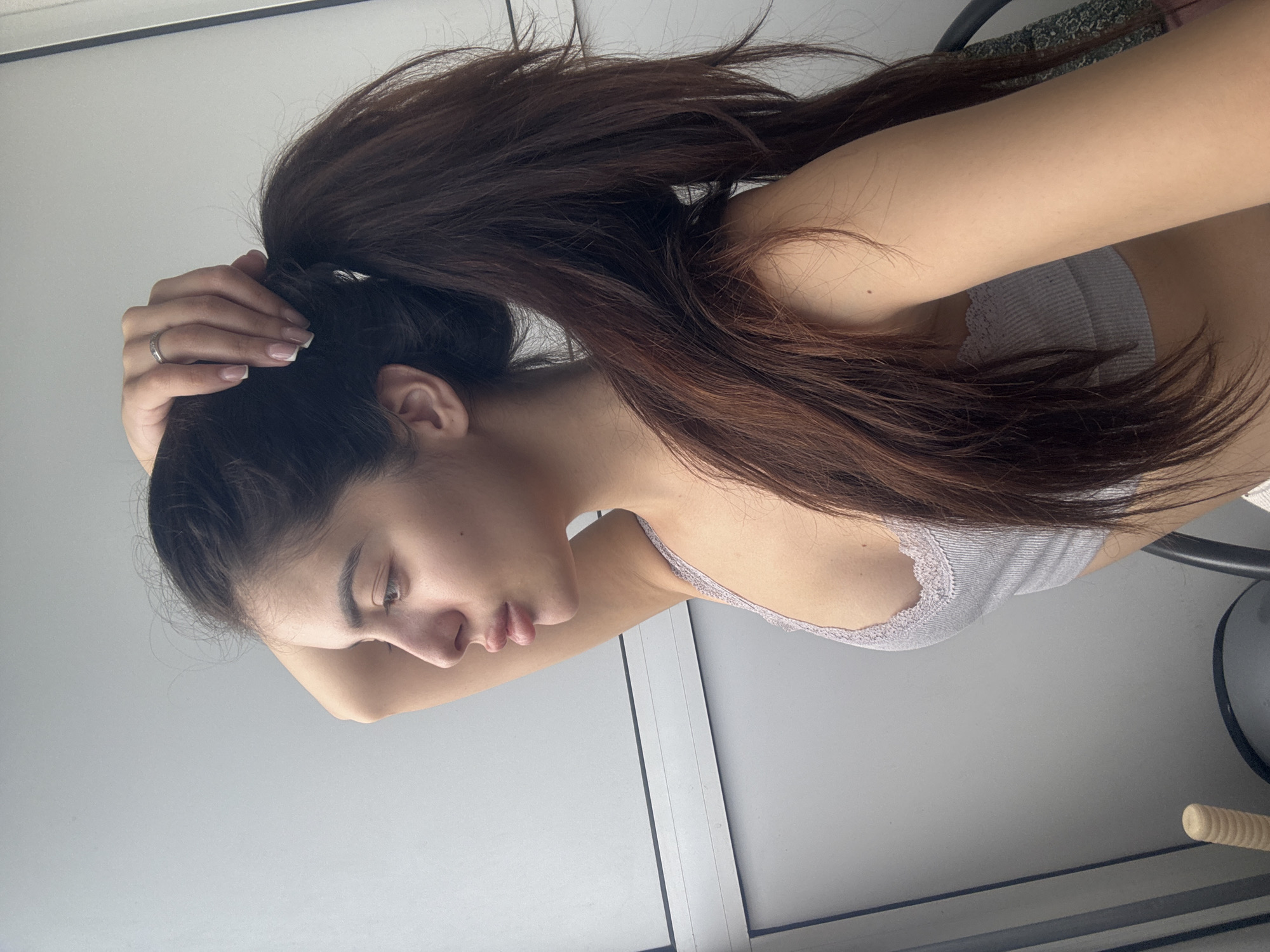 A young woman is depicted from the chest up, positioned indoors with a plain, light-colored wall or window as the background. She has long, dark brown hair styled in a high ponytail, which she is actively holding with her right hand at the back of her head. Her hand displays a silver-toned ring on her ring finger and her nails have a French manicure. She is wearing a light grey or lavender ribbed tank top or camisole with delicate white lace trim along the neckline. The woman's head is tilted downwards, and her eyes are cast low, suggesting a moment of focus or contemplation. The lighting is bright, indicating it is likely daytime. There is no visible text in the image. The location context for the image is Plovdiv, Bulgaria.