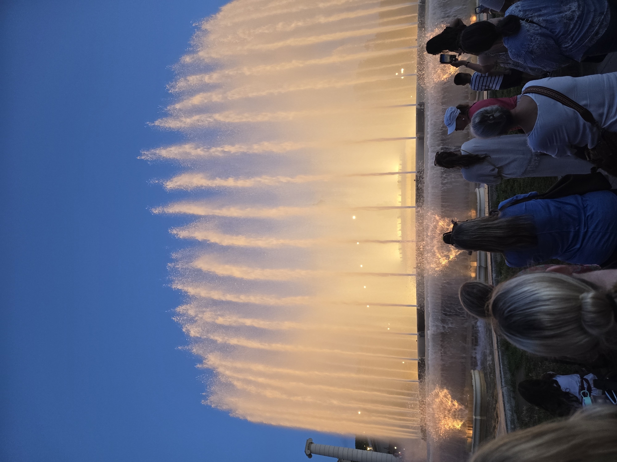The image depicts a view of the Magic Fountain of Montjuïc in Barcelona, Spain. A large, illuminated fountain is in the center of the frame, with numerous jets of water reaching high into the twilight sky. The water is lit with a warm, golden-orange glow. In the foreground, a crowd of people are gathered, facing the fountain and observing the spectacle. Some individuals are holding up phones, presumably to take pictures or videos. The sky above is a deep blue, indicating it is evening or early night. To the left of the fountain, a classical column is partially visible. The scene is set outdoors, with a grassy area in front of the fountain where some of the spectators are standing or sitting.