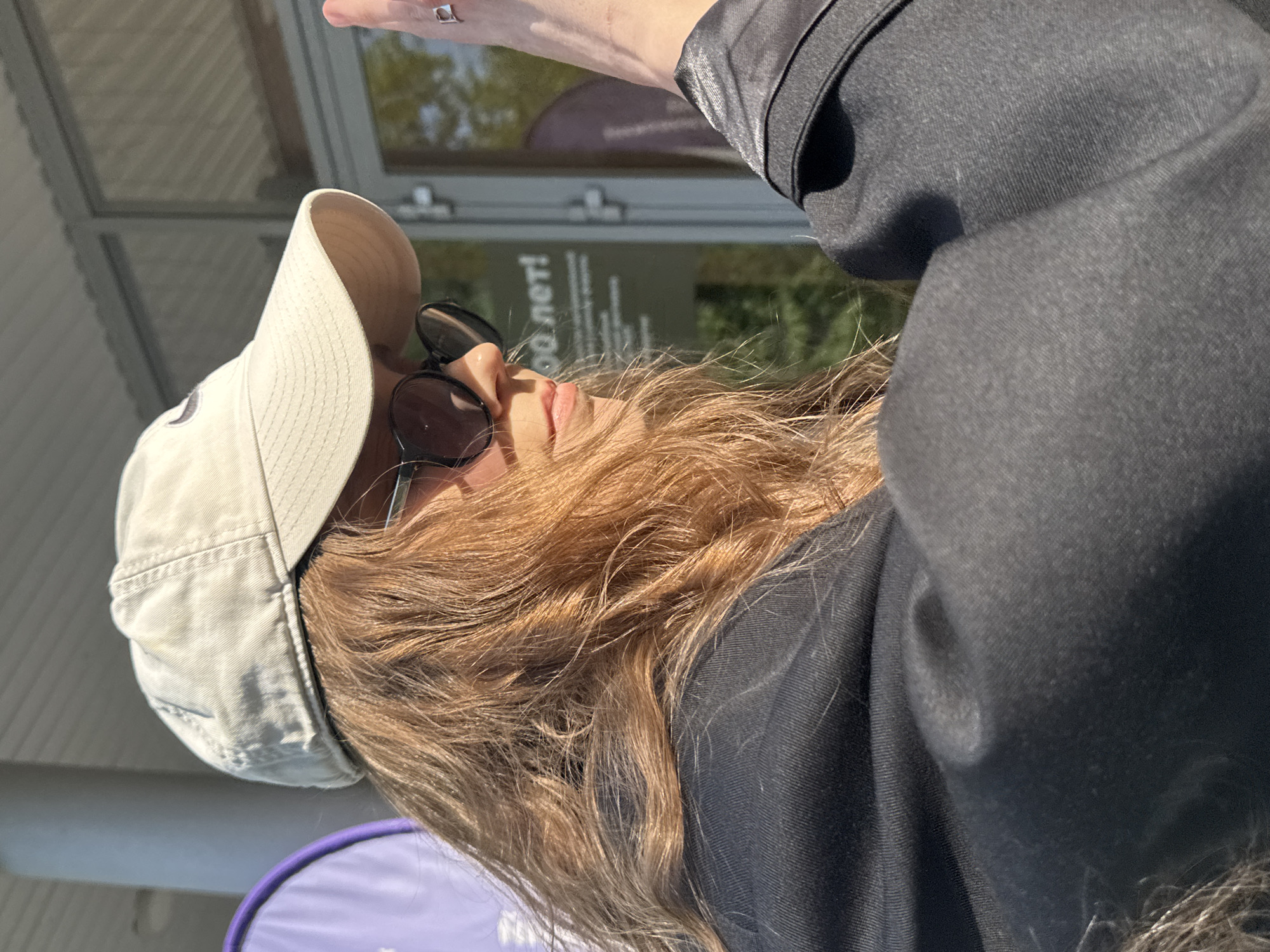A woman with long, light brown hair is wearing a light-colored baseball cap and dark sunglasses. She is wearing a dark-colored jacket. Her right hand is raised, potentially shielding her eyes from the sun. In the background is a building with the words "60 лет!" visible on a sign. The photo appears to be taken in Perm, Russia, based on the Cyrillic lettering on the sign.