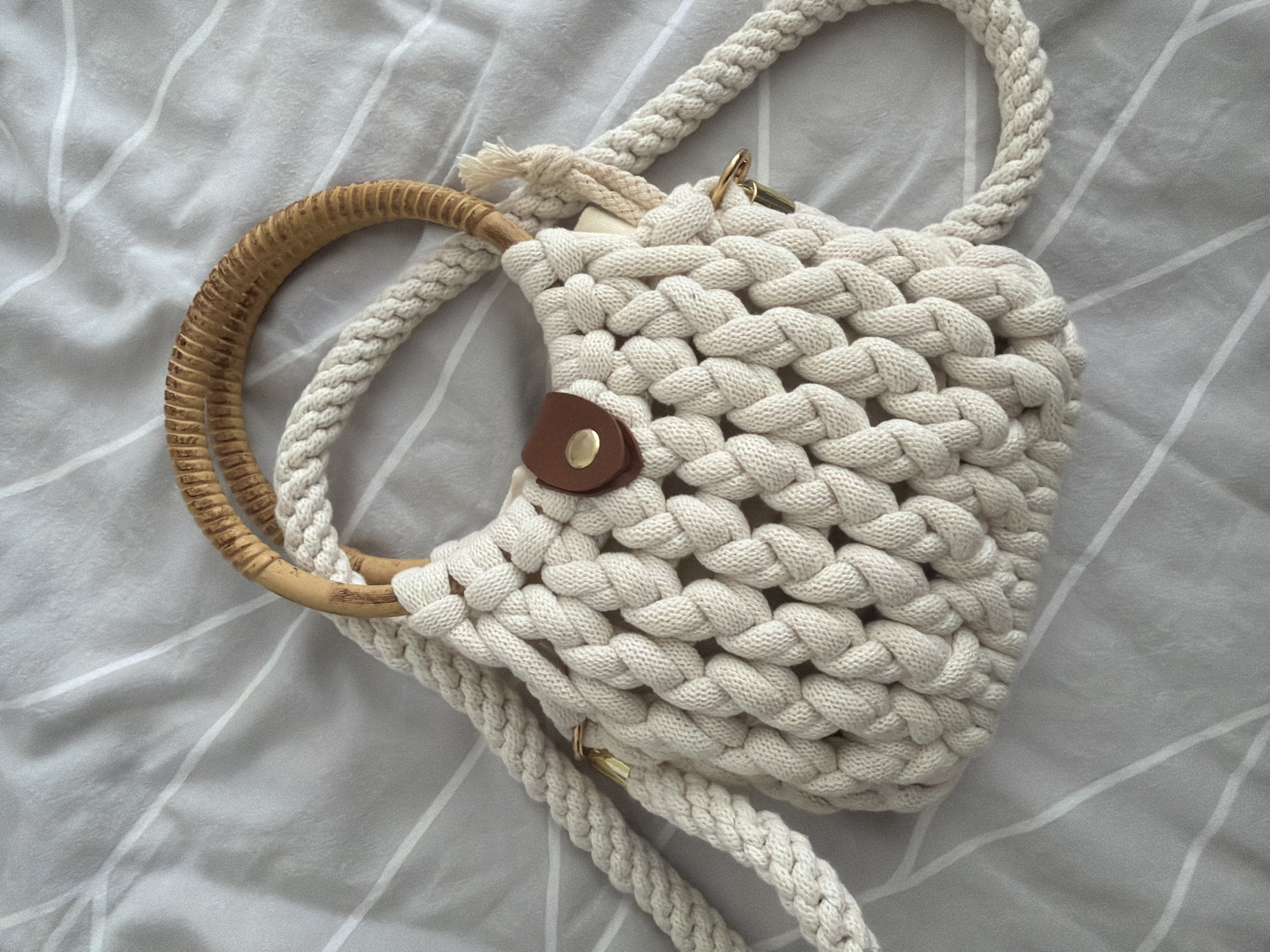 A cream-colored macrame handbag with a light brown woven handle and a brown leather snap closure lies on a gray and white striped surface, likely a bedspread. The handbag is intricately woven, featuring a detailed macrame pattern. Attached to the handbag are two braided cream-colored straps, one of which is draped over the side. The scene is captured from a top-down perspective. The image does not contain any information to determine a location in Pazardzhik, Bulgaria.