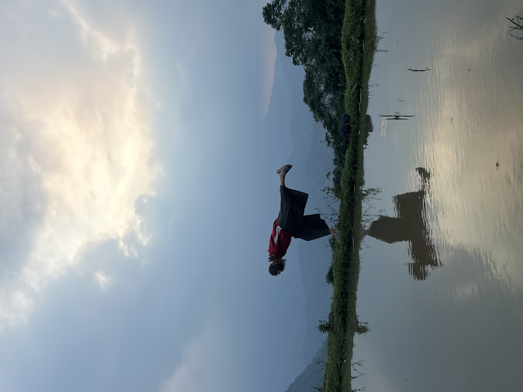 The image, correctly oriented, displays a serene natural landscape, likely in Nậm Dịch Commune, Vietnam, featuring a person in a dynamic pose.
The central subject is an individual, seen in profile, wearing a red long-sleeved top with white detailing on the sleeves and dark, wide-leg pants. They are performing an acrobatic or balancing pose, with one leg raised high behind them and their body leaning forward, appearing to be in motion or holding a stance. One grey athletic shoe is visible on the raised foot.
The setting consists of a tranquil body of water, possibly a rice paddy or a calm lake/river, that reflects the cloudy sky and distant elements. A narrow strip of land, covered with lush green vegetation including grass and small trees, separates the water from the background. In the distance, majestic mountains rise, appearing hazy and soft under the expansive sky. The sky is dominated by soft, diffused clouds in shades of white, light grey, and pale yellow, suggesting either early morning or late afternoon light. The water's surface is largely still, showing subtle ripples and reflections of the environment, including the person. A dark blue bag or object is partially visible on the ground near the water's edge.
The scene conveys a sense of peacefulness and an interaction between the human form and the natural environment. No visible text is present in the image.