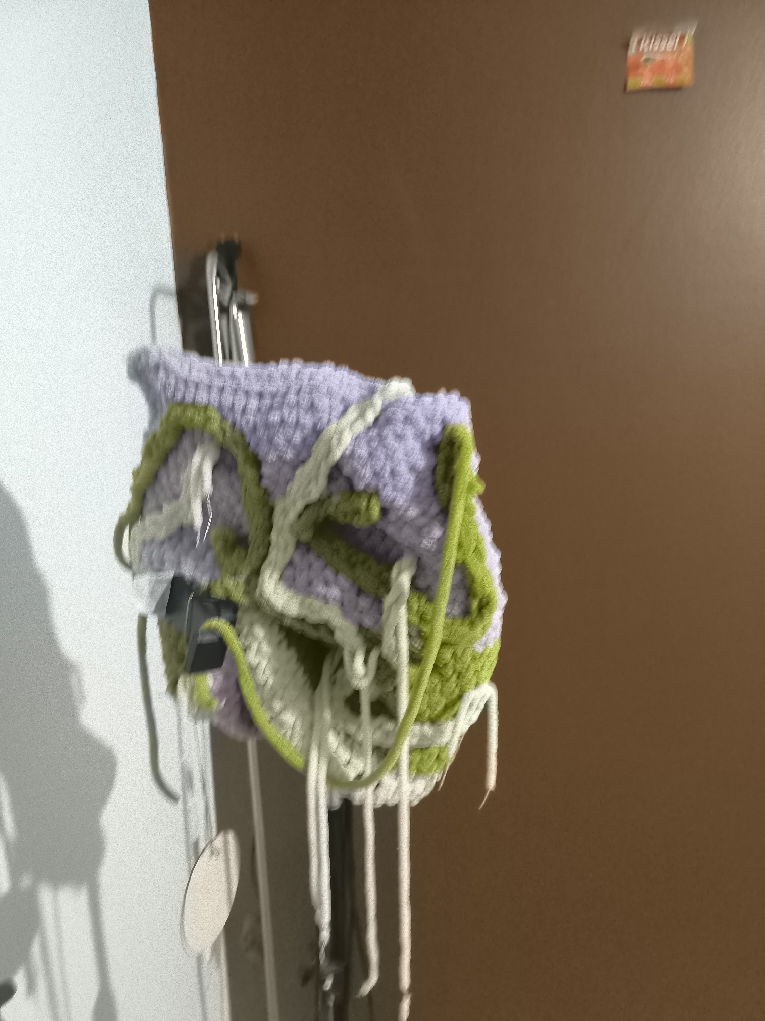 A knitted bag hangs on a metal hook against a brown door. The bag is primarily lavender with green and white yarn woven around it. Strands of white yarn hang down from the bottom. A white wall is visible to the left. A small red and yellow object is affixed to the door in the upper right corner. The bag is hanging at an undisclosed location and time. The city of Pélussin, France, is not indicated in this image.