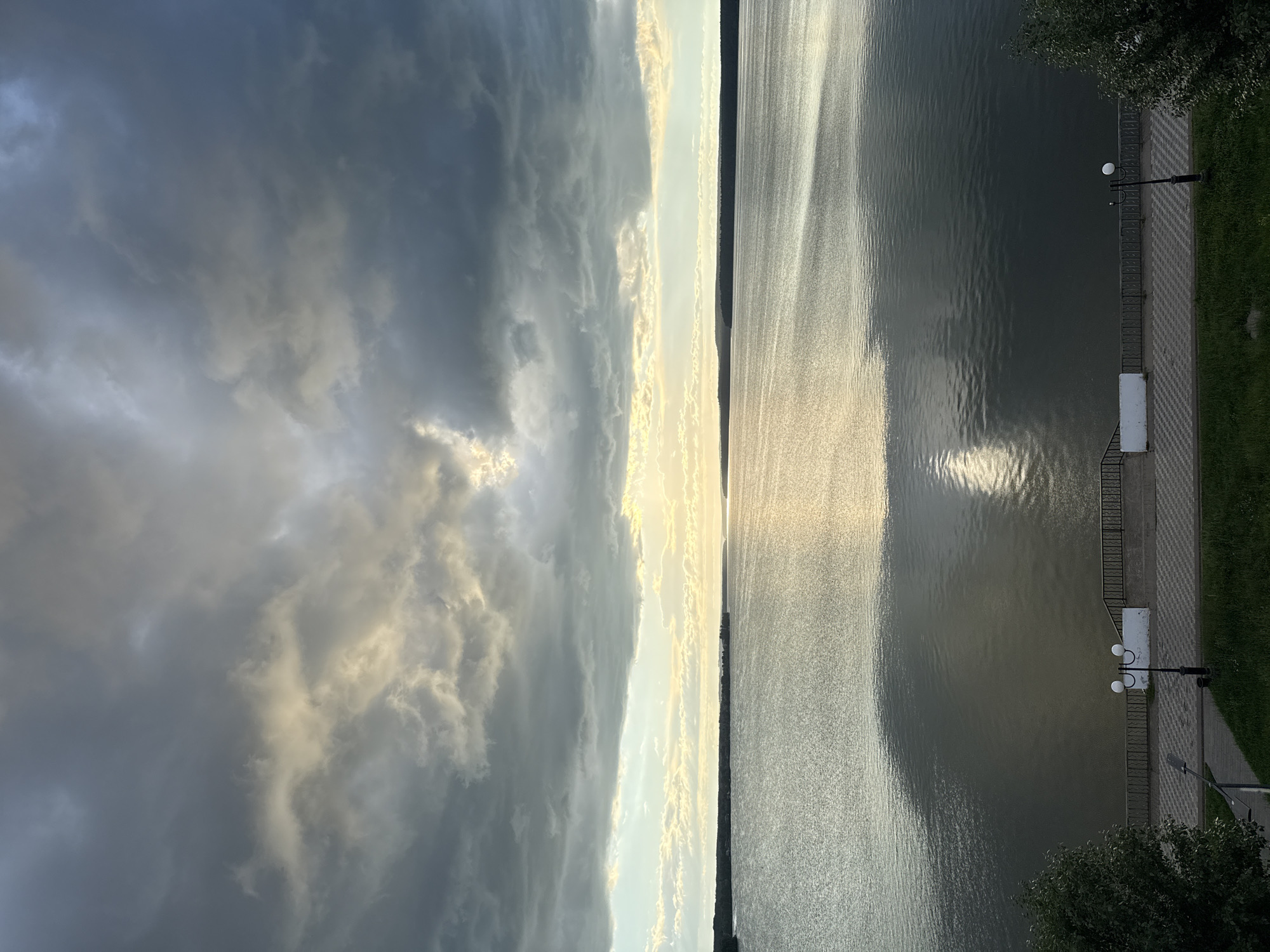 The image displays a view of a body of water, likely a river or lake, with a paved promenade alongside. The water is calm and reflects the light, creating shimmering patterns on its surface. The promenade is lined with a decorative fence and features streetlights, a bench, and a strip of green grass. Trees are visible on the right side of the frame, and in the distance, a faint outline of land can be seen. The sky is dramatic, with a mix of dark, ominous clouds and brighter areas where sunlight is breaking through, suggesting either a storm is passing or approaching. The light filtering through the clouds casts a soft glow on the scene, indicating it is either early morning or late afternoon. The overall mood is serene yet somewhat moody due to the contrasting light and shadow. The scene is set in Votkinsk, Russia. No people or text are visible in the image.
