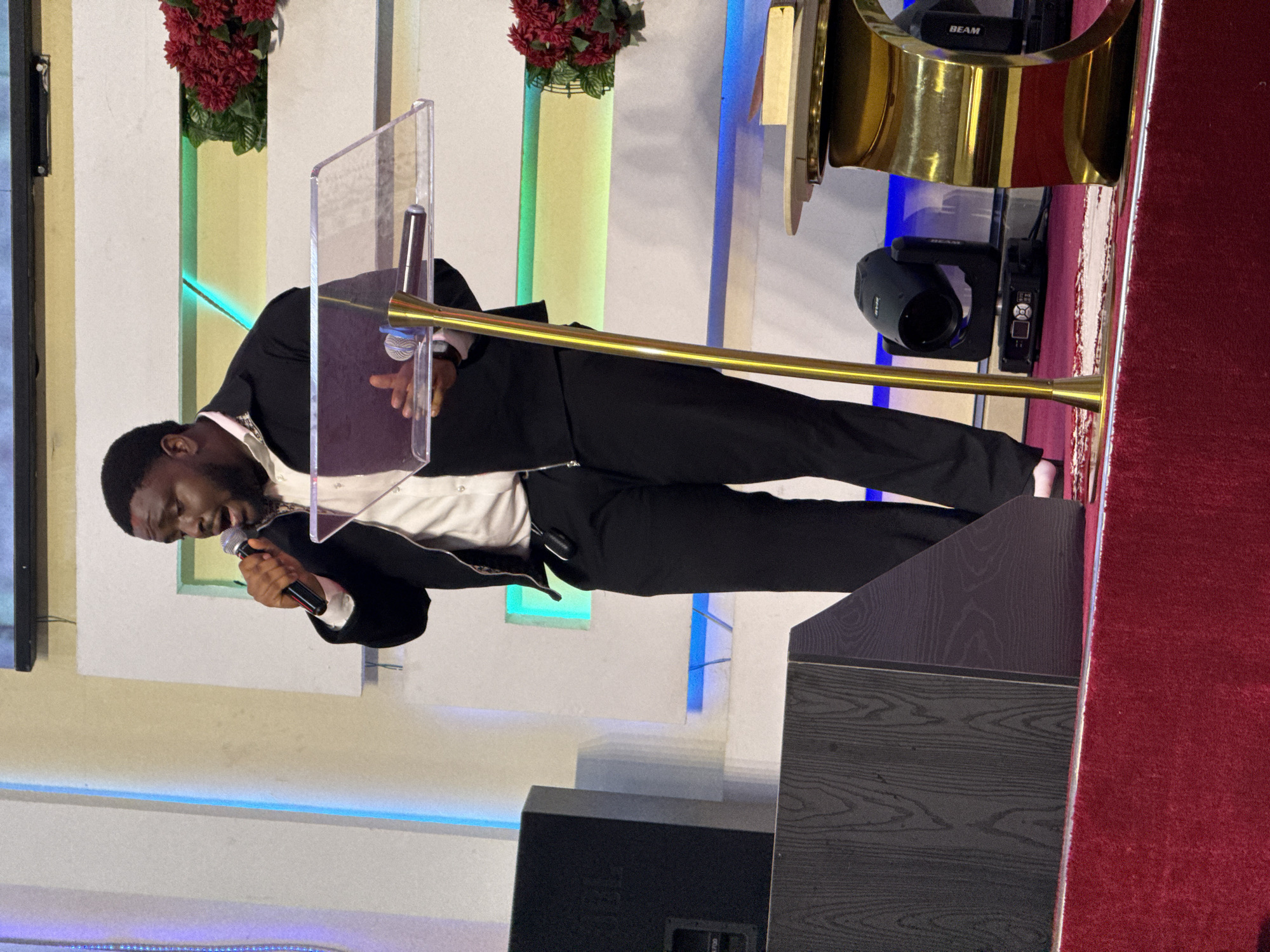 A man stands behind a clear lectern, holding a microphone and speaking into it. He is wearing a black suit jacket over a white shirt and black pants. The lectern sits on a gray wooden platform with a red carpet on the side. Behind the lectern, the wall is white with vertical green and blue stripes. Red flowers are visible at the top of the wall. Above the lectern, there is a gold structure with a beam light. A black speaker sits on the floor in front of the lectern. The photograph was taken at the location of Iddo Tudunwada, Nigeria.