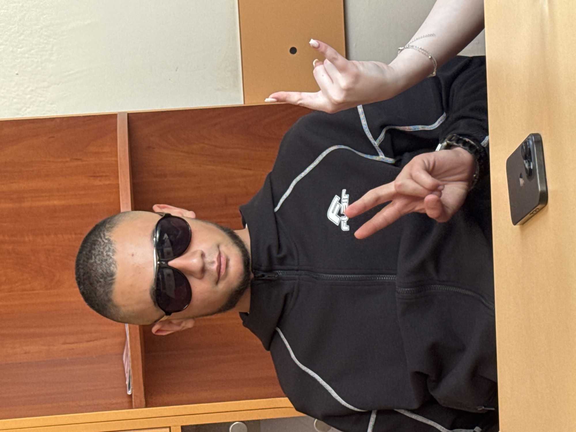 A person with short hair and sunglasses is shown in the image. They're wearing a black zip-up jacket with white trim. Their right hand is raised, making a peace sign. Another person's hand is also visible above the first, fingers splayed, fingernails painted white. There is a phone leaning against a wooden surface to the right of the frame. The background features wooden shelves and a tan wall. The image was taken indoors. There is no information about the location or the city of Elhovo, Bulgaria.