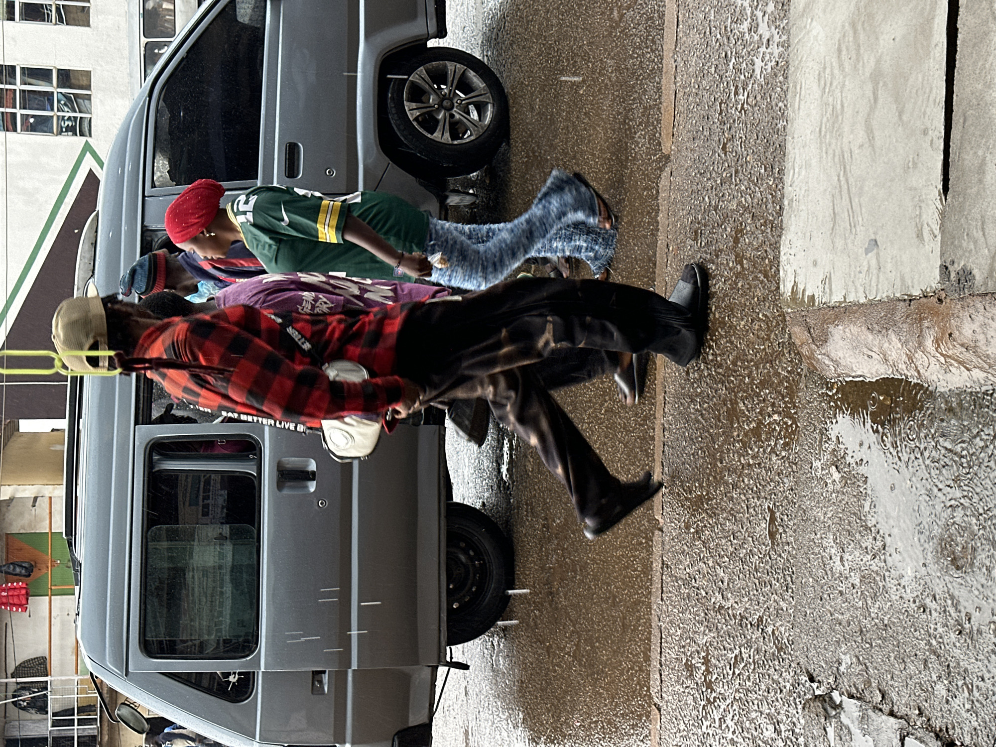 The image depicts a rainy street scene in Bukuru, Nigeria, with several individuals walking alongside a grey minivan. The ground is wet and shows puddles, indicating recent or ongoing rainfall.
In the foreground, a person wearing a red and black plaid shirt and dark pants is walking away from the camera, with their left leg extended. They are carrying a white bag. To their left, another person in a purple shirt is partially visible. Further to the right, a woman in a green and yellow jersey with the number 12 on it, and a red headscarf, is walking. Her legs are clad in light blue, textured pants. Behind her, another person wearing a blue and red hat is also walking.
The grey minivan is positioned on the right side of the image, with its front half visible. The windows are tinted. Several people are partially visible inside the van. The environment suggests an urban setting with buildings in the background, some of which have window grids. Clothes are hanging on a rack outside one of the buildings. The overall impression is of a candid street photograph taken during inclement weather.