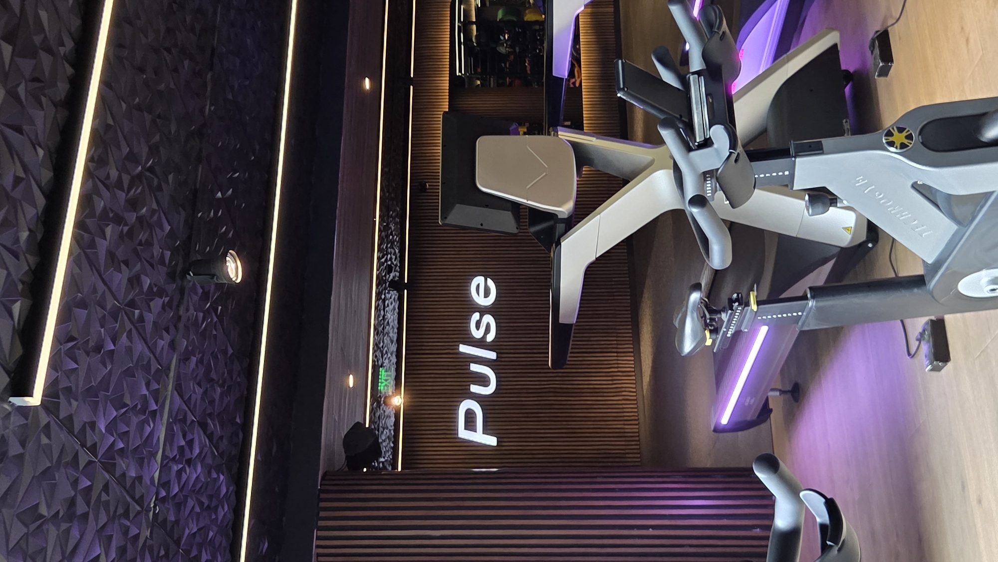 The image shows a Pulse fitness center. The "Pulse" logo is prominently displayed on a wood-paneled wall. An exercise bike, facing left, is next to the wall with the Pulse logo. The environment includes dark textured walls with accent lighting. A small spotlight illuminates the textured wall. The location in the city of Unknown, Unknown is not possible as this is not mentioned.