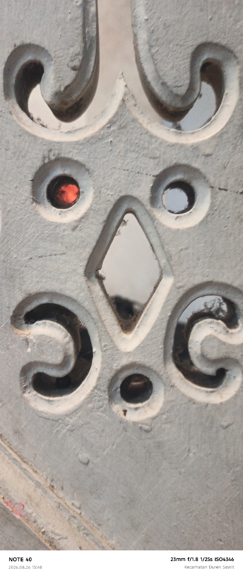 The image is a close-up, macro photograph of a decorative, textured, grey surface with carved details. The surface appears to be outdoors, possibly part of a building's facade or an ornamental element. Several circular indentations are visible, one of which contains a small, red, reflective object. Another indentation is diamond-shaped and contains a reflective surface, likely a mirror or polished material, showing a distorted reflection. The overall texture of the surface is rough and granular, with visible imperfections and slight damage. The lighting suggests it is daytime, casting subtle shadows within the carved areas. The bottom of the image contains text metadata: "NOTE 40 2026.08.26 13:48" and "23mm f/1.8 1/25s ISO4346 Kecamatan Duren Sawit". Based on the location information "Kecamatan Duren Sawit", the scene is located in the Duren Sawit subdistrict, East Jakarta, Indonesia. There are no people or animals visible in the frame.