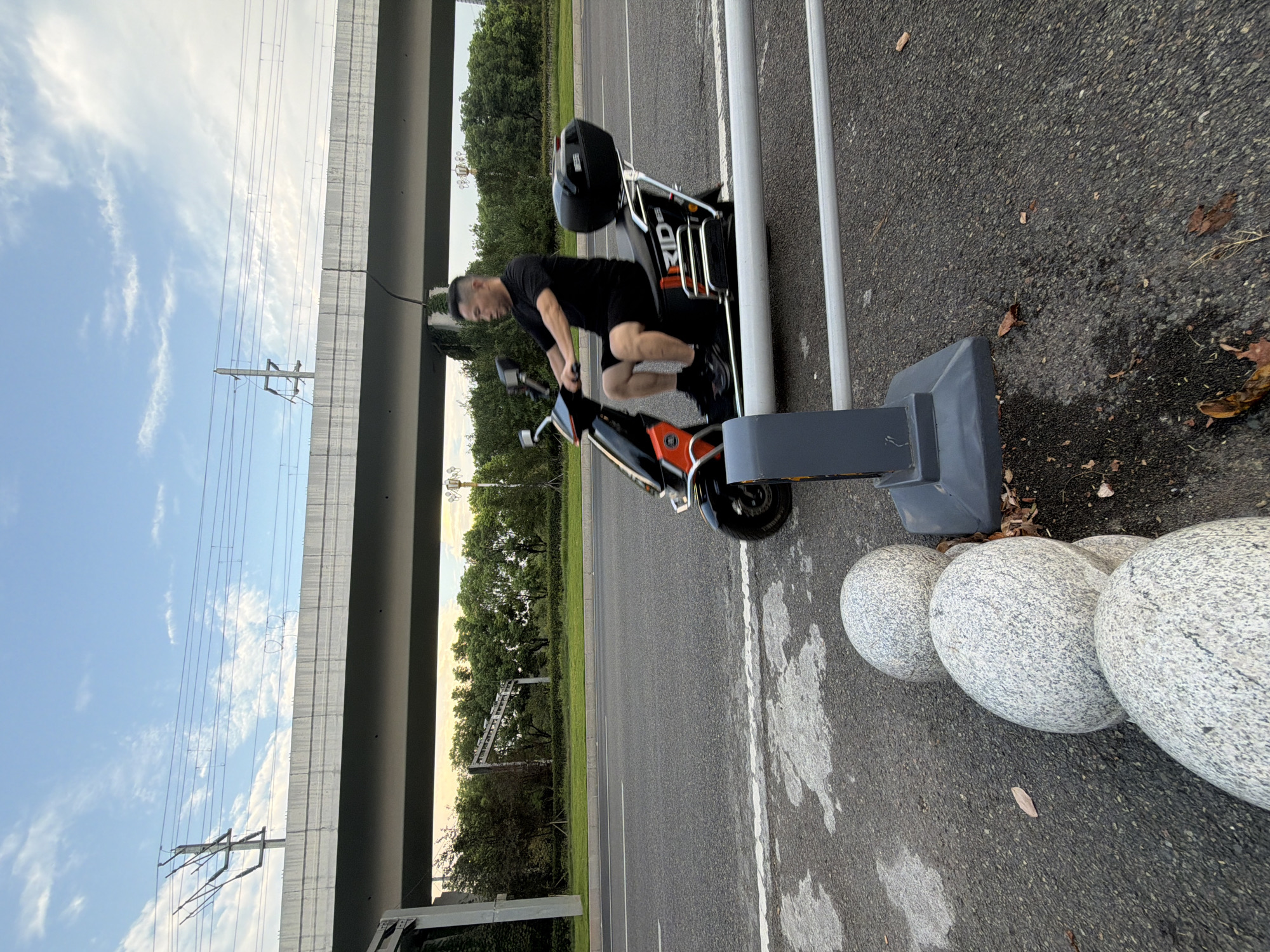 A middle-aged Asian man in a black t-shirt and black shorts rides a black and orange electric scooter along a paved road. He is looking to his left. The scooter has a black storage box on the back and the word "RID" is visible on its side. The man is wearing black socks and black shoes.
In the background, a large concrete overpass runs horizontally across the image. Above the overpass, power lines are visible against a blue sky with scattered white clouds. Trees and greenery line the sides of the road and beneath the overpass.
In the foreground, several large, rounded grey stones are arranged in a line on the side of the road. A grey metal railing with a white pipe runs across the foreground, partially obscuring the scooter. The road surface is asphalt with some wet patches and fallen leaves. The scene appears to be set in Jiangjin, China.