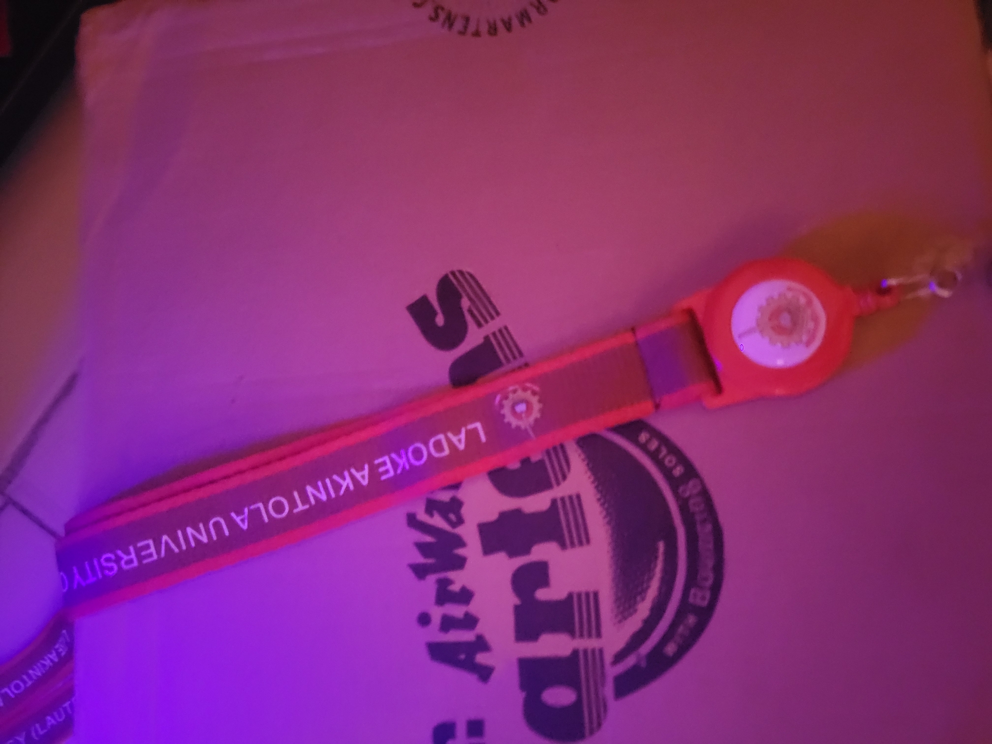 A close-up, high-angle, overhead shot displays a lanyard with text on a pinkish background, likely a box. The lanyard is orange with red trim and text in white that reads "LADOKE AKINTOLA UNIVERSITY". Below this text, there are two small circular logos with gears inside. At the bottom of the lanyard, a retractable reel is attached with a silver clip. The background appears to be a textured surface. The lighting in the image is dim and casts a purple and pink hue.