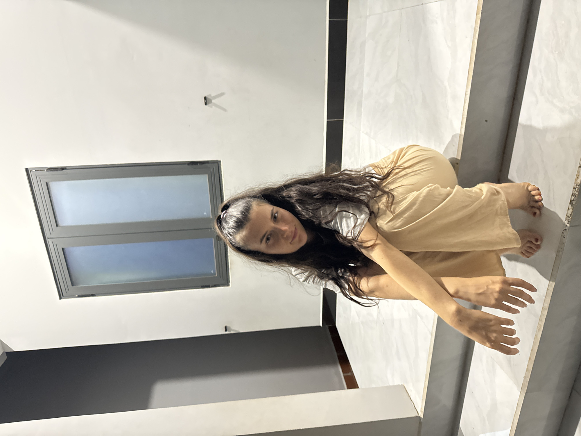 A woman with long, dark hair, wearing a light-colored top and tan pants, is crouching on a marble staircase. She is looking at the camera. The location appears to be indoors, with a window visible in the background. The specific location is Mũi Né, Vietnam.