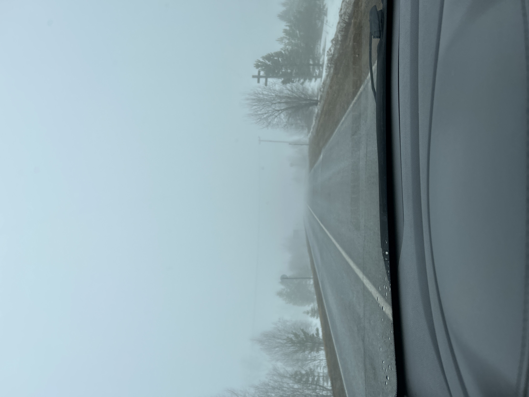 The image, shot from inside a vehicle, depicts a foggy or snowy rural road in what appears to be Уральское, Russia. The road stretches into the distance, obscured by heavy fog or snow. A white line marks the edge of the road. Bare trees line both sides of the road, and power poles can be seen. The vehicle's windshield and a portion of the dashboard are visible in the foreground. The wiper blade sits to the side of the windshield, with small droplets along the windshield indicating moisture.