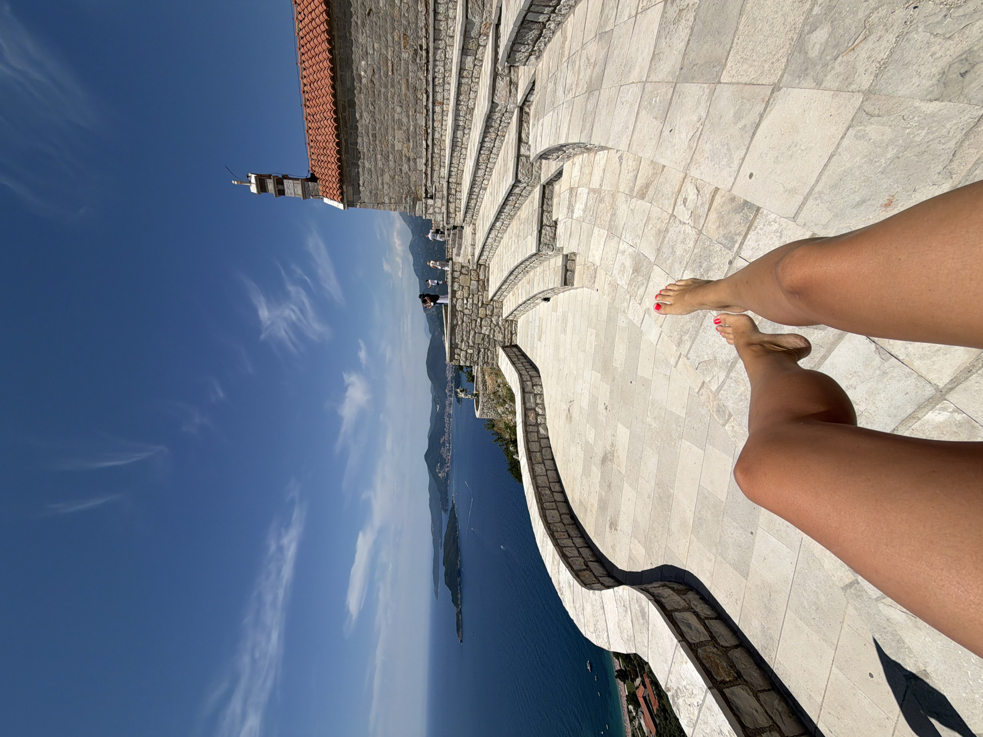 The image depicts a person's legs and feet in the foreground, resting on a paved patio area. The person's toenails are painted red. The patio is made of large, light-colored square stones and is bordered by a low stone wall. Beyond the wall, there is a panoramic view of the Adriatic Sea and the coastline. In the distance, the island of Sveti Stefan is visible, along with the mainland coast of Montenegro. There are mountains in the background, and a clear blue sky with wispy white clouds.
To the right of the patio, there is a stone structure with a red-tiled roof and a small bell tower. In front of this structure, there are tiered stone steps that form an amphitheater-like seating area. Several people are visible on the steps and near the stone structure, some appearing to be tourists.
The overall scene suggests a scenic overlook in Sveti Stefan, Montenegro, possibly a historical site or a viewpoint. The lighting indicates it is daytime, likely sunny.