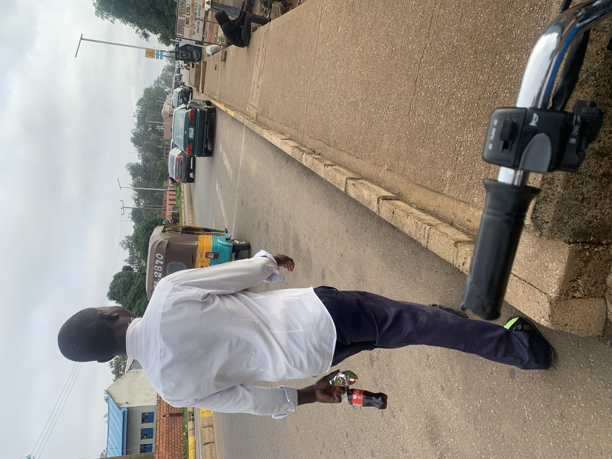 This image, captured in Barakin Lerawi, Nigeria, depicts a daytime street scene under an overcast sky. In the foreground, a person wearing a white long-sleeved shirt and dark trousers is seen from behind, walking along the edge of a paved road. They are holding a small Coca-Cola bottle and a packaged item in their left hand. To the right, handlebars with visible controls, including "TURN" and "CHOKE" labels, indicate the photo was likely taken from a motorcycle or scooter.
The road features several vehicles, including a dark green sedan and another dark car in what appears to be a line of traffic. Further down, a yellow and blue auto-rickshaw, or Keke Napep, is visible with the number "2870" on its side. On the right, a concrete sidewalk runs parallel to the road, with a person in dark clothing seated upon it in the distance.
The background shows various buildings, including one with a distinct blue roof, and numerous trees. Utility poles and lines crisscross the sky. A prominent sign on a building reads "LOBMAK PHARMA (NIG. LTD.)," with additional text below it stating "NAF GATE SHOPPING PLAZA" and "ABBATTOIR RAYFIELD." The scene portrays a typical urban or semi-urban environment with ongoing activity.