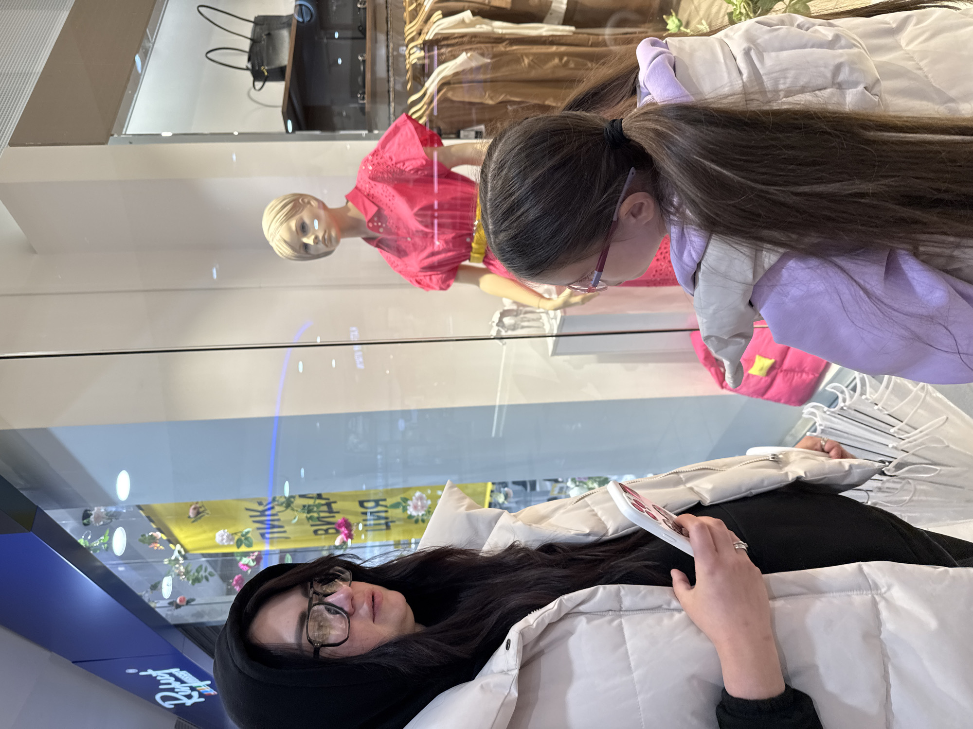 A low-angle, medium shot captures a mother and daughter looking into a shop window. The mother is lying down, wearing a black hoodie and glasses. She has long, dark hair and a cream-colored puffy jacket. She is holding a white phone in her left hand, and a ring is visible on her left ring finger.
The daughter, positioned above the mother, is also wearing glasses and has long, dark hair pulled back in a ponytail. She is wearing a cream-colored jacket with a purple hood. Both are looking at the items displayed inside the shop window.
Inside the window, a mannequin wearing a pink top and yellow skirt is on display, along with clothing racks. The lighting inside is bright, reflecting on the glass. The overall environment suggests an indoor shopping mall.
The location is the city of Unknown, Unknown.