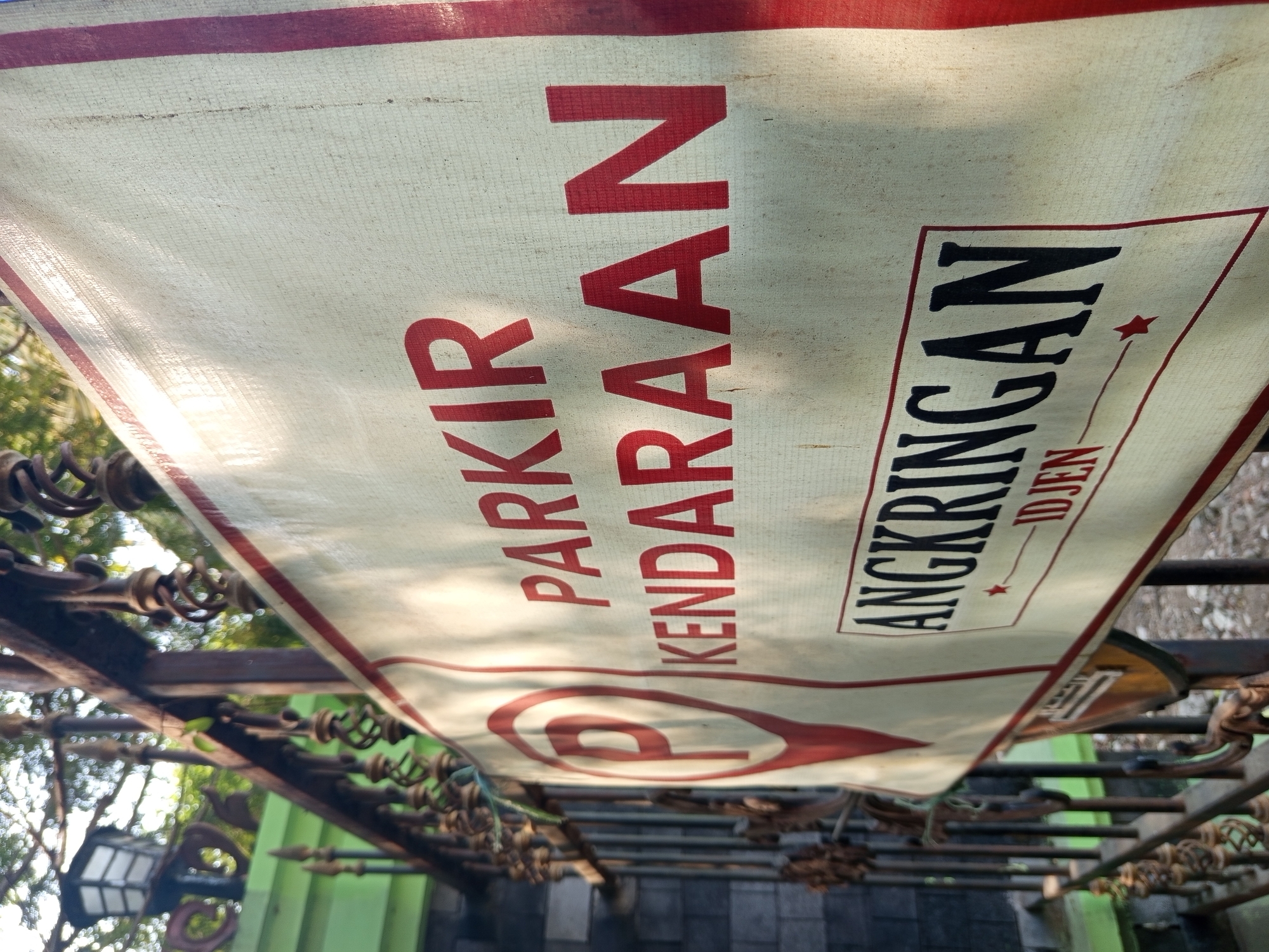 The image shows a close-up of a white banner with red and black text, hanging on a dark metal fence. The banner reads "PARKIR KENDARAAN" (Vehicle Parking) in large red letters, and below that, "ANGKRINGAN" (a type of food stall or eatery) and "IDJEN" in smaller black letters. A red parking symbol, a "P" inside a red pin icon, is visible on the left side of the banner. The banner is slightly creased and appears to be made of a fabric material. The fence is ornate with spiral metalwork. Behind the fence, a green wall and a black brick wall are visible, along with foliage and a street lamp. The lighting suggests it is daytime, with dappled sunlight falling on the banner. The text "IDJEN" in the context of Malang, Indonesia, likely refers to the "Djenggring" or "Djengger" area, which is a tourist spot and a natural landmark in the city, known for its cultural significance and often associated with local eateries. Therefore, the sign likely indicates a parking area for vehicles visiting "Angkringan Idjen" in Malang.