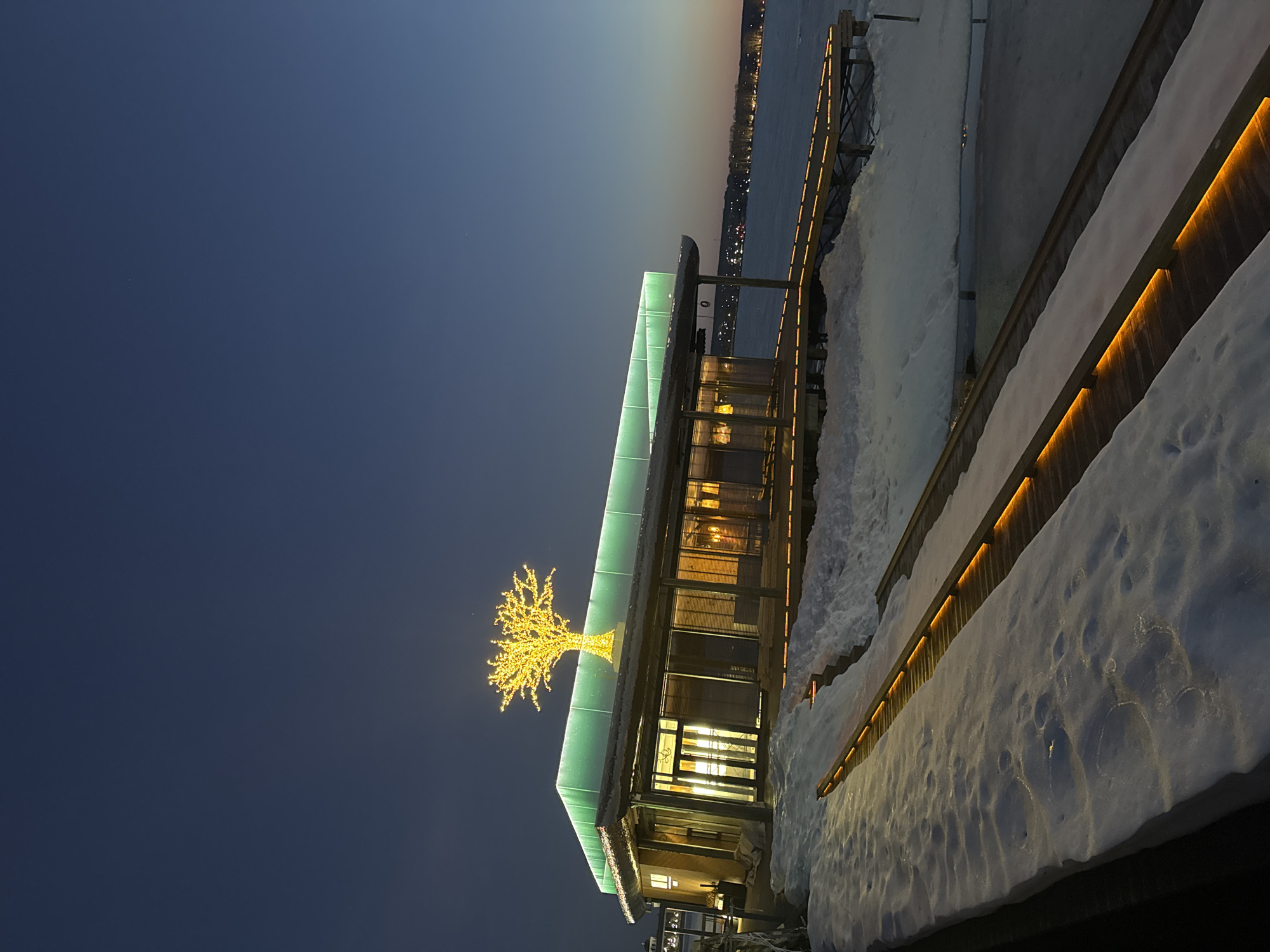 A building with a lighted tree on top is partially covered in snow near a body of water. The location is Votkinsk, Russia. The building has a green roof and appears to have glass walls, with interior lights visible. A wooden railing is also covered in snow, with a light strip running along its length. The sky is dark blue to the left and fades to orange near the horizon.