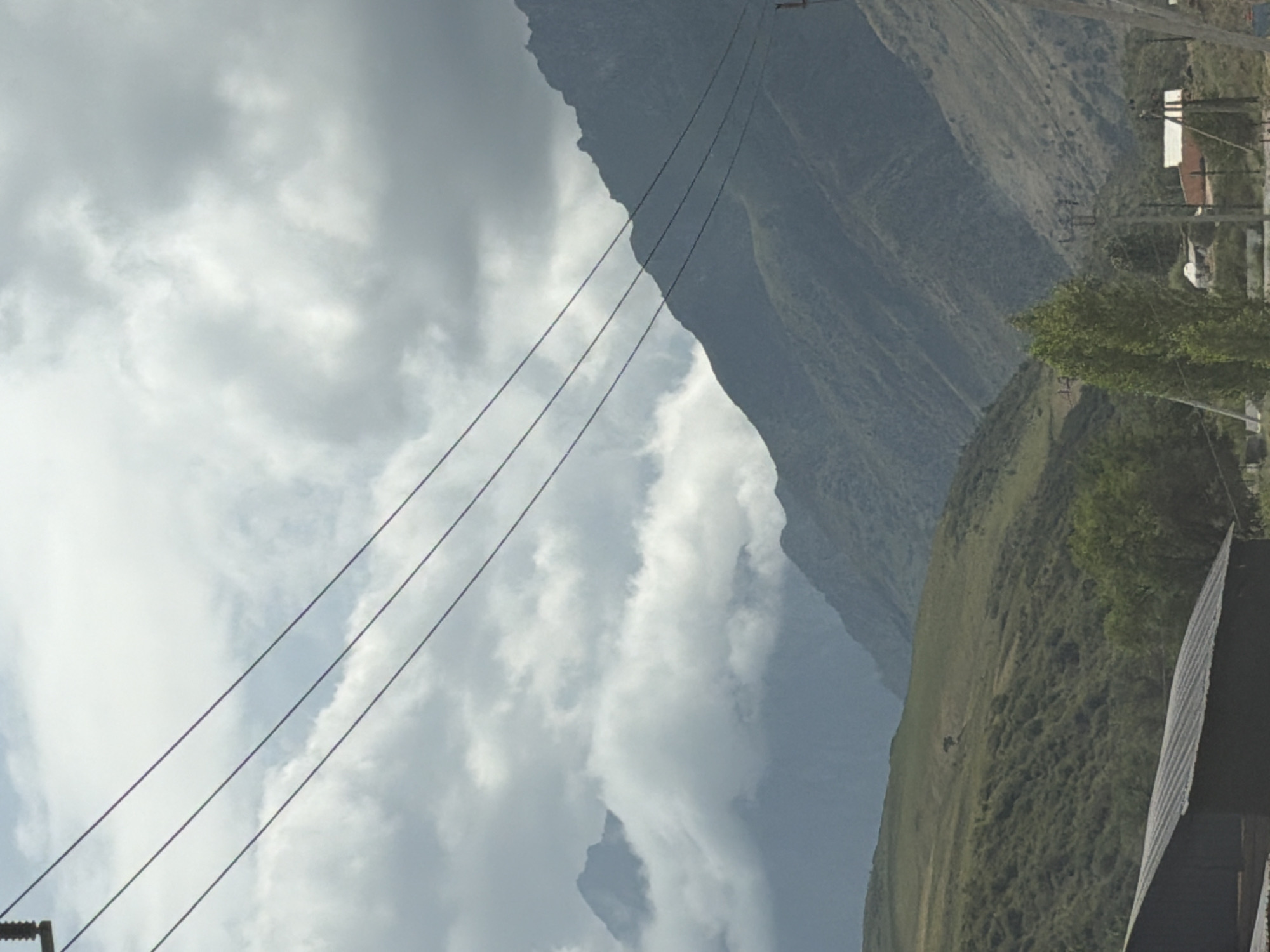 The image displays a mountainous landscape under a cloudy sky. In the foreground, a dark, sloped roof structure is visible on the left. Several power lines stretch across the frame from left to right. The middle ground features rolling green hills with scattered trees and a solitary tree in the distance. In the background, majestic mountains dominate the scene, with some peaks obscured by thick clouds. A few buildings and power poles are sparsely scattered throughout the landscape. The overall impression is one of a remote, natural setting.